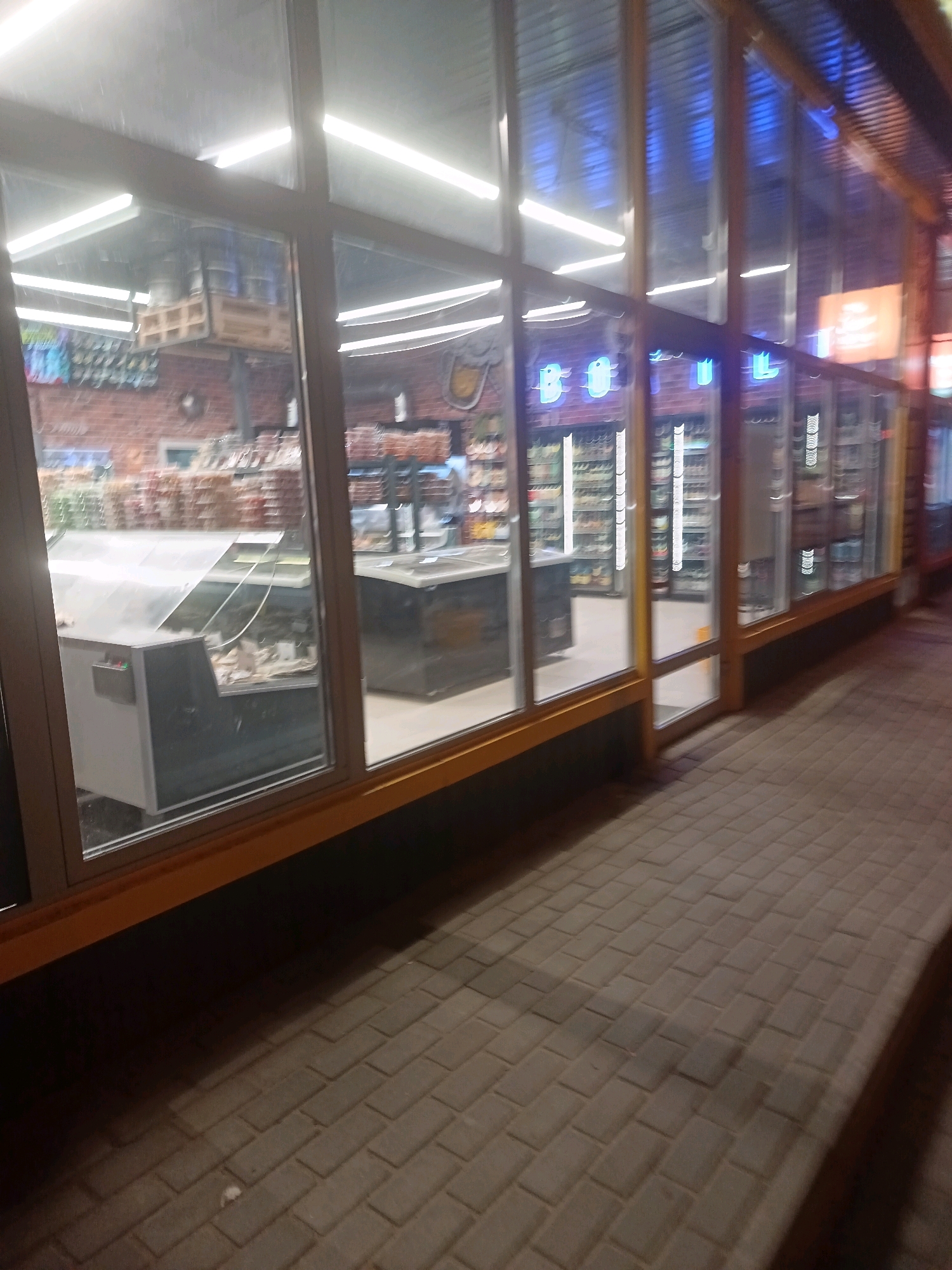The image shows the exterior of a shop at night, viewed from the sidewalk. The shop has large glass windows that reveal the interior, which appears to be a convenience store or small market. Inside, there are shelves stocked with various products, refrigerators, and display cases. The lighting inside is bright, with fluorescent lights illuminating the merchandise. There are no people visible in the image, either inside or outside the shop. The sidewalk in front of the shop is paved with rectangular bricks. The overall impression is that of a typical urban storefront at night. The location is identified as Astrakhan, Russia.
