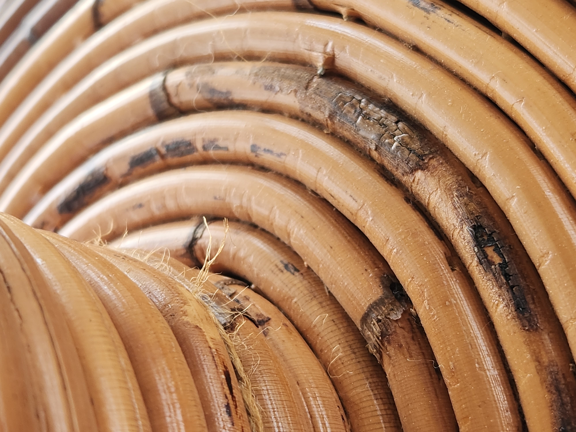 The image is a close-up, abstract shot of a woven rattan object. The texture of the rattan is the dominant feature, showcasing its natural fibers and a glossy finish. Some areas of the rattan appear darker and more weathered, with visible cracks and frayed strands, suggesting age or wear. The object is cylindrical, with the weaving forming concentric, curved lines that recede into the background. The lighting highlights the curved surfaces and creates subtle shadows. The image does not contain any identifiable elements that would allow for a specific location to be determined, nor does it depict any people, actions, or interactions.