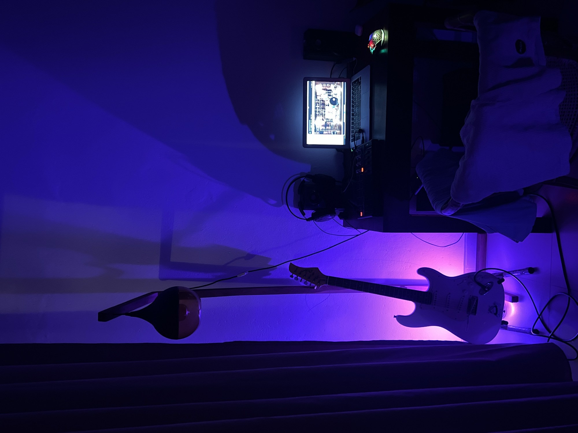 The image shows a room interior with purple lighting. On the right, there's a desk area with a laptop, a monitor displaying a screen interface, headphones, a speaker, and miscellaneous electronic equipment. A chair with a white cushion is also present. Two stringed instruments are mounted on the wall: a teardrop-shaped tanbur and a white electric guitar. The bottom of the frame shows a dark, wavy curtain.
The image was taken in a room, presumably at night. The location is Goviyapana, Sri Lanka.