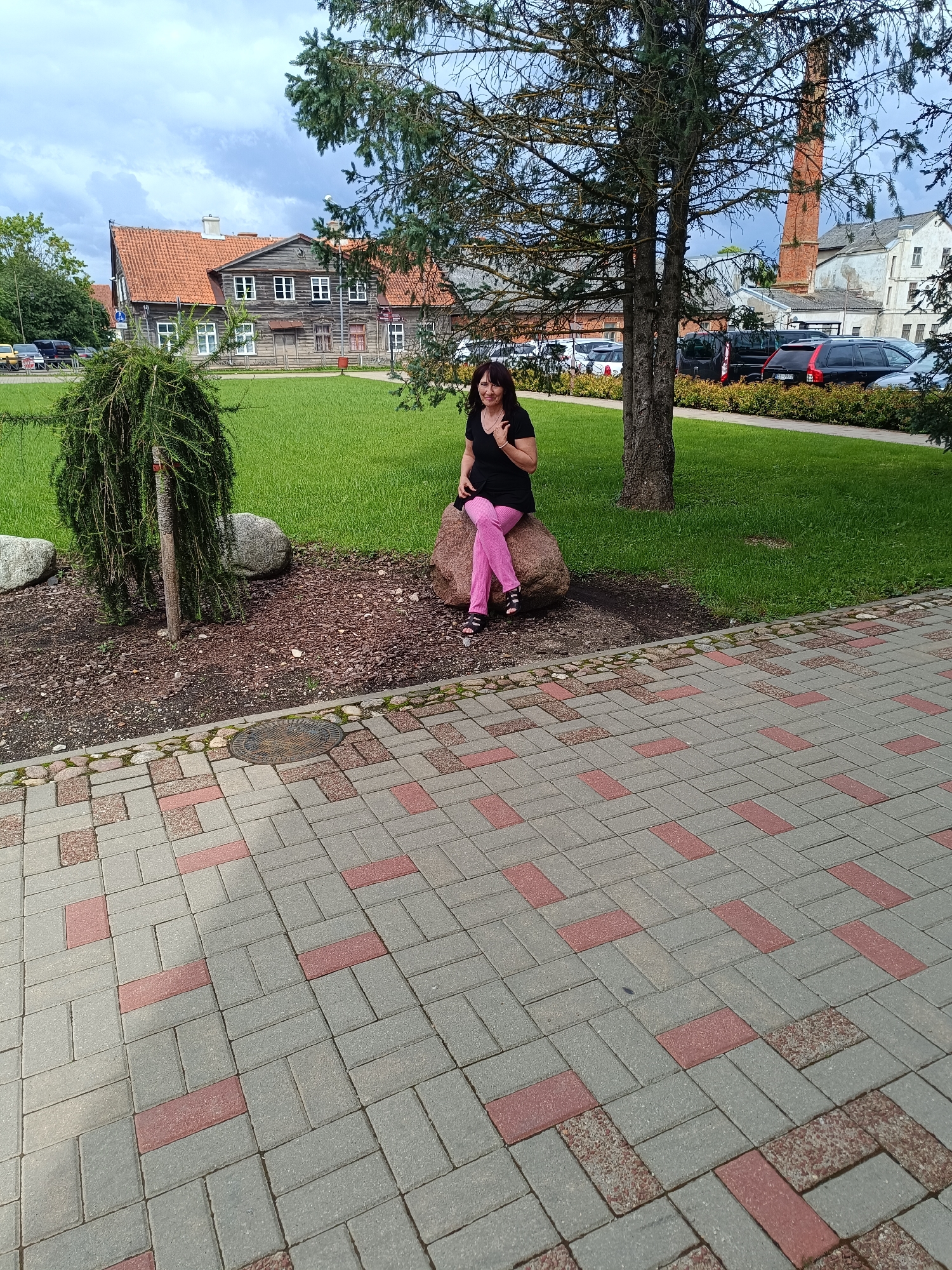 A woman is sitting on a large rock in a grassy area with trees and buildings in the background. She is wearing a black top and pink pants. The woman is looking towards the camera and smiling. The background shows a traditional wooden building with a red tile roof, a brick chimney, and several cars parked in a lot. The foreground is a paved walkway made of gray and red interlocking bricks. This scene is likely in Kuldīga, Latvia, given the architectural style of the buildings and the cobblestone paving.