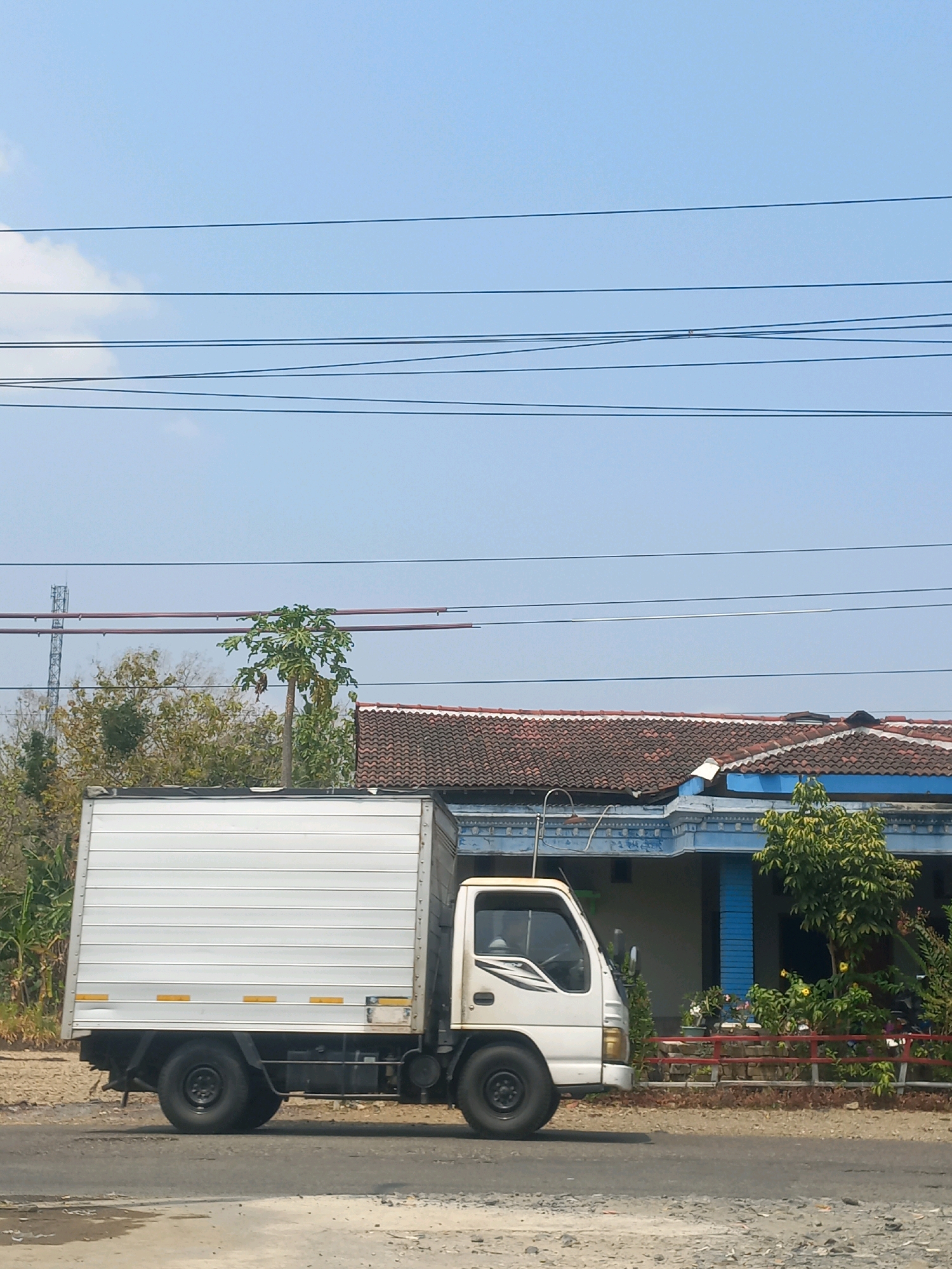 A white box truck is parked on the side of a road in Baturetno, Indonesia. The truck has a large, rectangular cargo area with horizontal silver paneling and yellow reflectors. The cab of the truck is white with black trim. In the background, a single-story house with a red tiled roof and blue trim is visible. A papaya tree stands to the left of the house. Several power lines are strung across the bright blue sky. The road appears to be asphalt with a gravel shoulder. There are no people visible in the image.