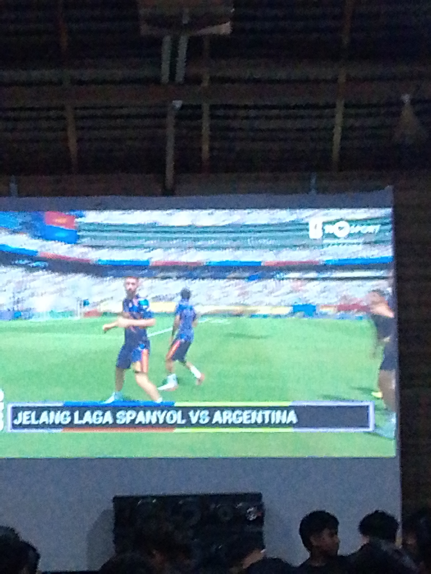 The image shows a large screen displaying a soccer game. Two players in blue and red uniforms are on the field. Text on the screen reads "JELANG LAGA SPANYOL VS ARGENTINA," which translates to "Before the Spain vs. Argentina match." The screen is positioned in front of an audience, with the heads and shoulders of several people visible in the foreground. The background appears to be a thatched roof structure. The scene is likely taking place in Meunasahmee, Indonesia, based on the provided location.