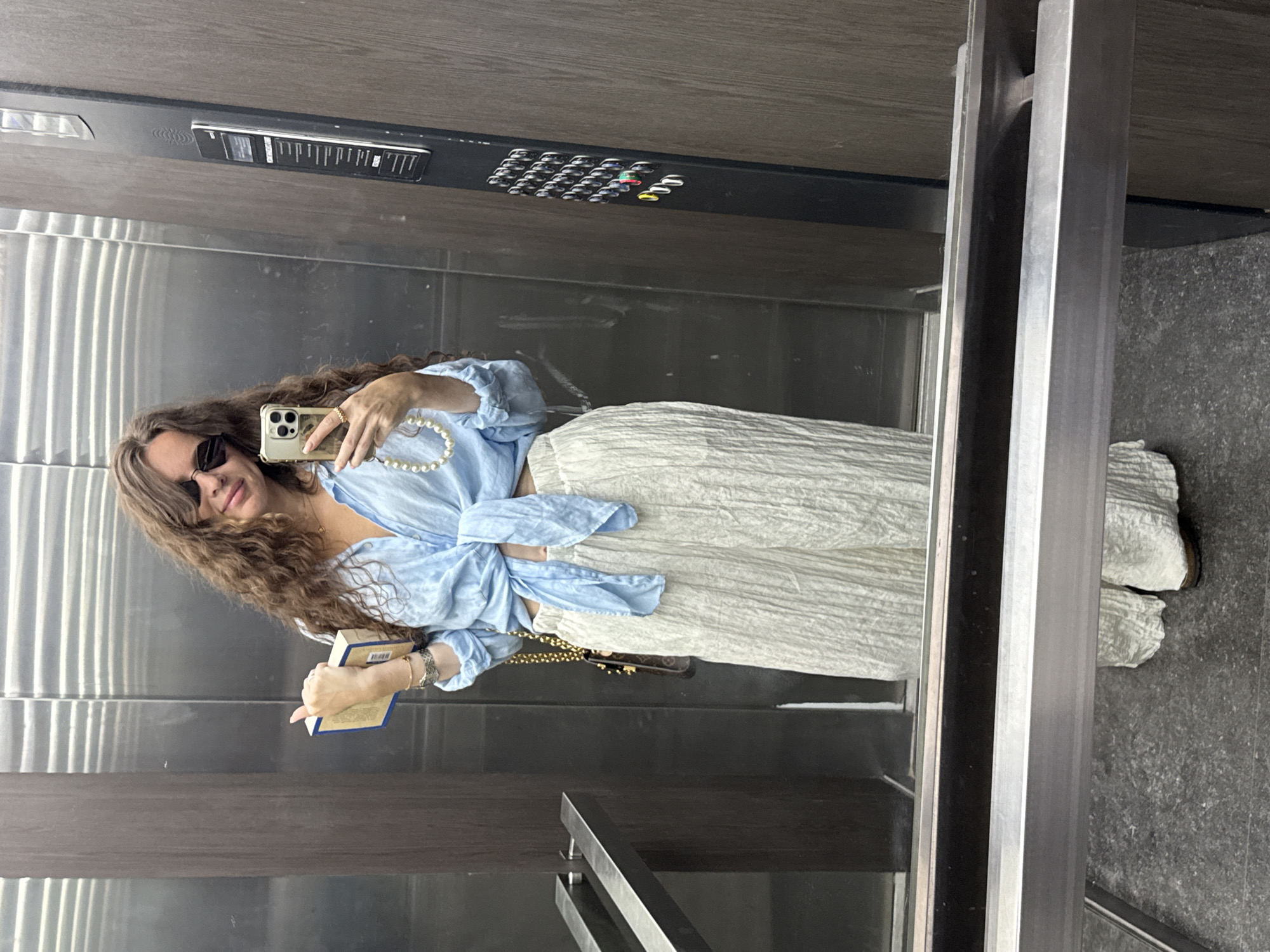 A woman with long, curly brown hair, wearing sunglasses, a light blue tied-front shirt, and off-white wide-leg pants, stands in an elevator. She is holding a book in her left hand and her smartphone, with a pearl phone strap, in her right hand, as if taking a selfie. The elevator has stainless steel walls and doors, a wooden panel on the right side with an elevator control panel, and a brushed metal handrail. The floor is dark gray. The scene appears to be within a modern building. There is no specific visual information to indicate the location is Moscow, Russia.