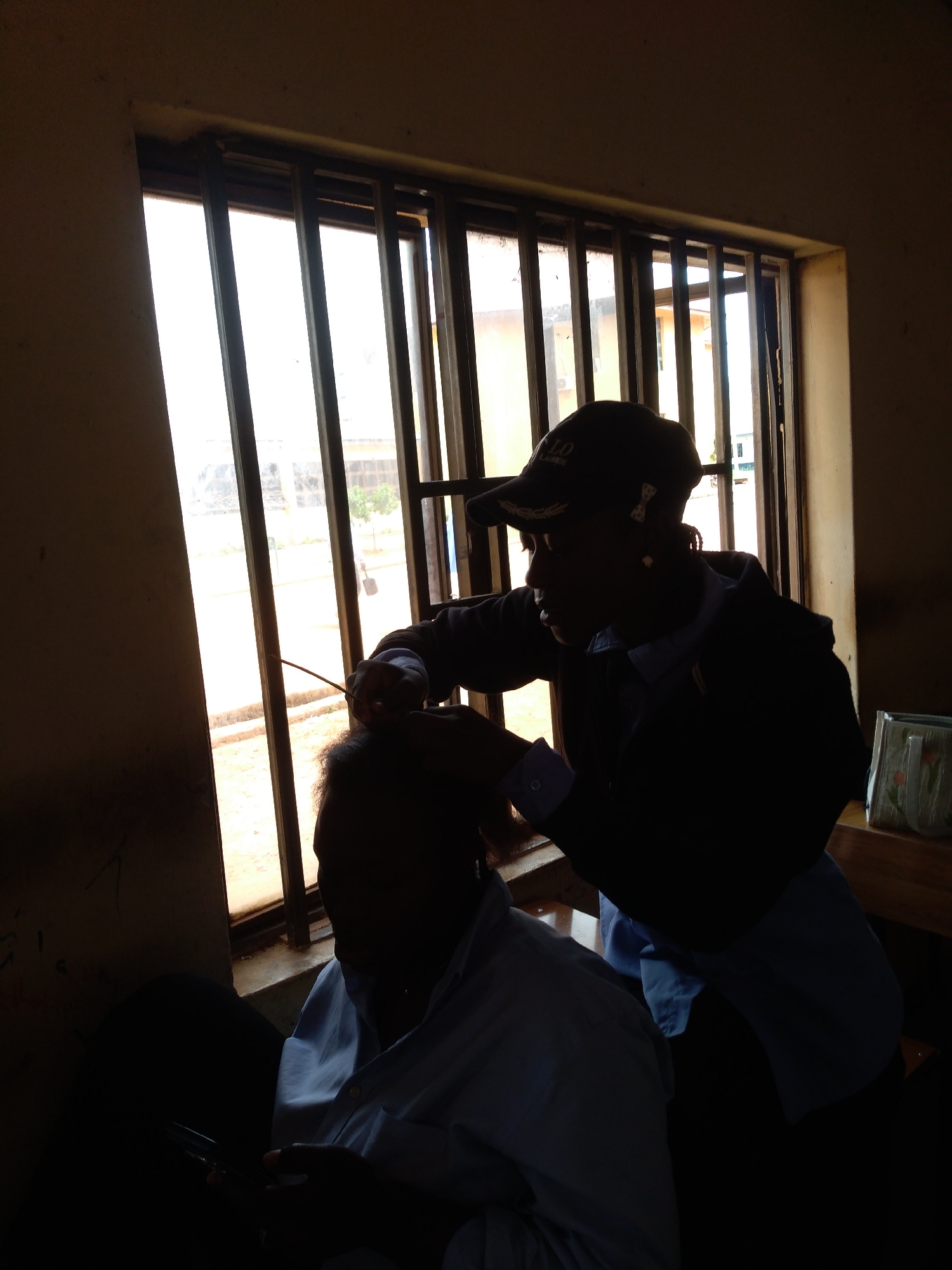 In a dimly lit room, two individuals are positioned near a window with vertical bars. The person on the right, wearing a baseball cap and a dark jacket over a collared shirt, is leaning forward and appears to be interacting with the hair of the person on the left. The person on the left is seated and facing away from the viewer, their back and the back of their head visible. They are wearing a light-colored collared shirt and are holding a dark object, possibly a phone, in their hands. The background visible through the window shows a bright, overcast sky and hints of buildings and foliage, suggesting an outdoor environment. A patterned bag is visible on a wooden surface to the right of the frame. The overall scene is silhouetted, obscuring fine details but indicating a close-up interaction. The location is identified as Jos, Nigeria.