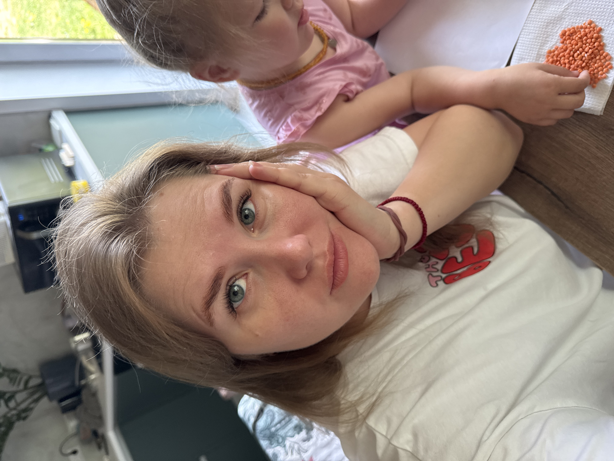 A young adult female with light hair and blue eyes is taking a selfie. She is wearing a white t-shirt with a red design on it. Her left hand is resting on her face. A small child wearing a pink shirt and a necklace is behind her. They are sitting at a wooden table. There is a white piece of paper on the table, and an orange cluster of small spheres on a white napkin. The setting appears to be indoors in a residential area. The location is Ферма, Russia.