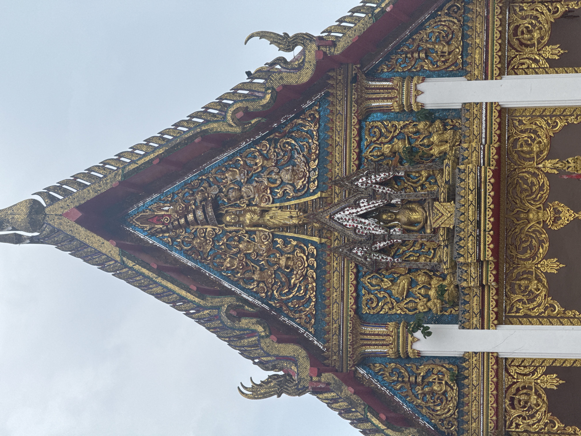 This is a close-up, eye-level, outdoor, vertical shot of the ornate gable of a Buddhist temple in Chalong, Thailand. The temple's facade is heavily decorated with intricate gold-colored relief work against a teal background, featuring depictions of figures and swirling patterns. The triangular gable is topped with layered, decorative elements in gold, red, and dark colors, resembling scales or layered finials. Flanking the central gable are curved, decorative finials resembling mythical creatures or stylized dragon heads, also in gold and red. Two white pillars are visible on either side of the lower part of the facade, partially obscuring the detailed gold work beneath the gable. A few green leaves are visible growing from crevices in the architectural details, suggesting the presence of vegetation. The sky above is overcast and light gray, providing a neutral backdrop. There are no people visible in the image. The overall impression is one of elaborate craftsmanship and religious artistry.