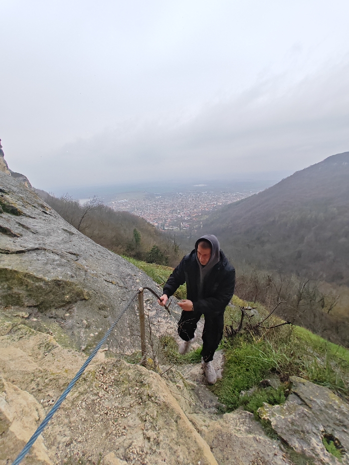 The image depicts a steep, rocky mountain trail overlooking the city of Shumen, Bulgaria, under an overcast sky. In the foreground and mid-ground, an adult male, wearing a dark hooded jacket over a grey hoodie, black trousers, and light-colored sneakers, navigates the uneven terrain. He grips a curved metal support post of a safety cable railing with his right hand, while his left hand holds a small, light-colored object, possibly a mobile device. The blue-sheathed steel cable railing runs diagonally upwards from the lower-left. The path consists of light brown and grey rock formations, loose gravel, and patches of green grass and small, bare shrubs. In the mid-distance, the city of Shumen is visible in a valley, characterized by numerous buildings with reddish roofs. Beyond the city, a large, indistinct body of water is faintly discernible. The surrounding mountain slopes are covered with dense, mostly bare trees. The sky is uniformly grey and cloudy.