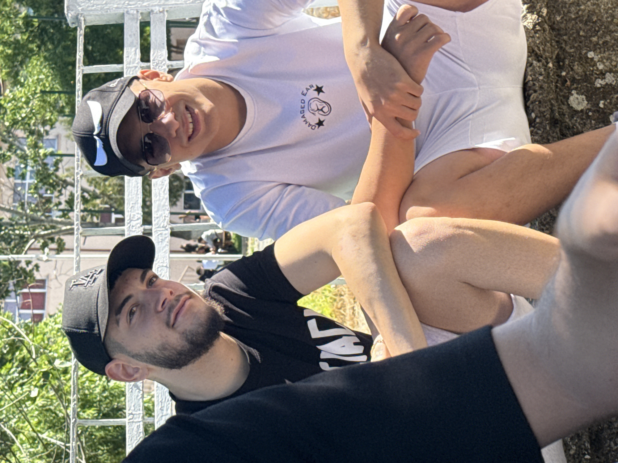 Two young men are seated outdoors on a white metal railing or structure, possibly a bridge or elevated walkway. The man at the top, oriented to the left, wears a white t-shirt with a logo, sunglasses, and a black baseball cap. He is smiling. The man at the bottom wears a black t-shirt and a black baseball cap with a logo, and he has a beard. His gaze is directed upwards. It is daytime with bright sunlight. The environment includes trees and buildings in the background, suggesting an urban or park setting. Due to the lack of detail in the image, it is difficult to identify the exact location in the city of Elhovo, Bulgaria.