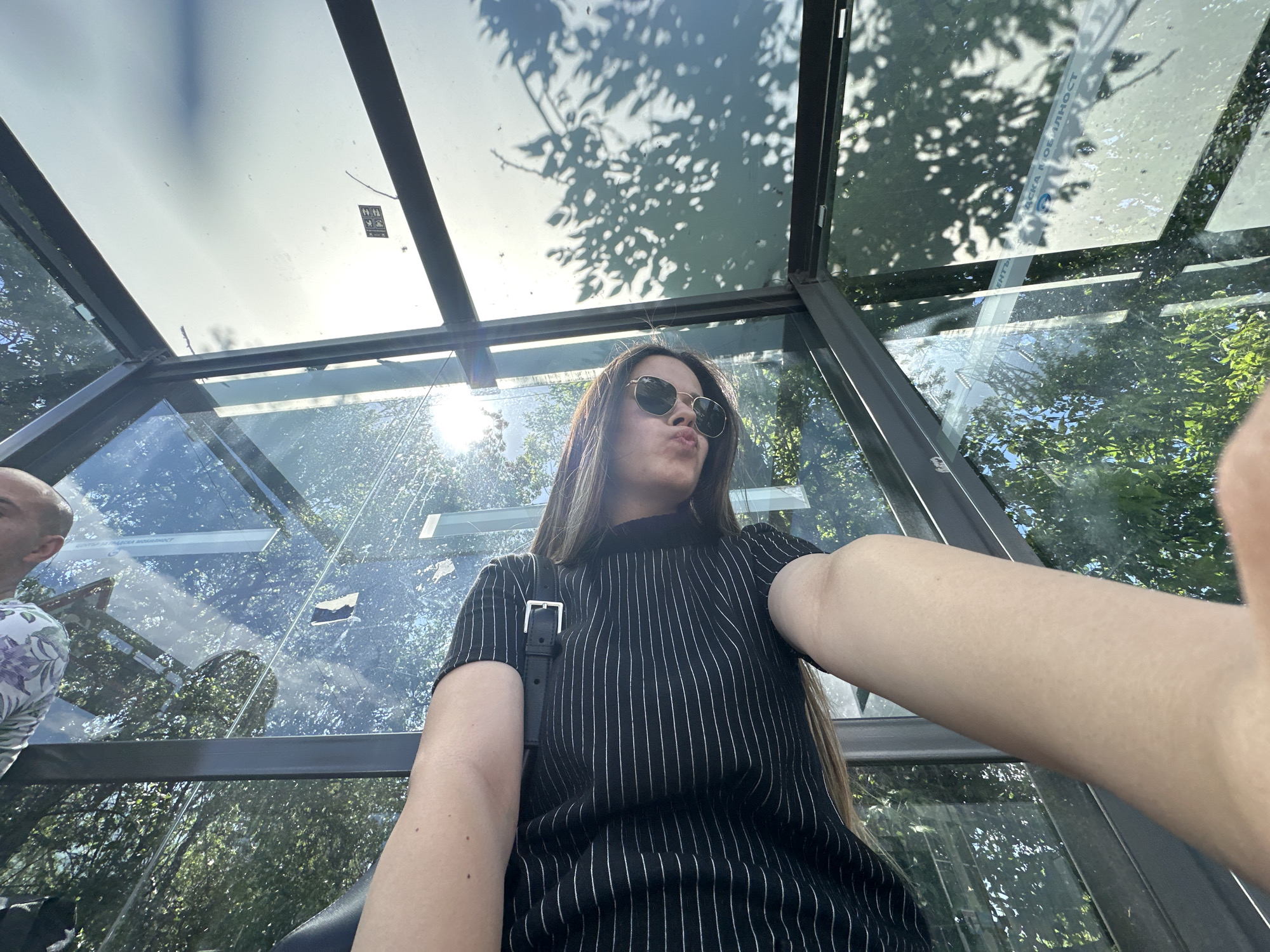 A low-angle shot shows a woman with long brown hair and sunglasses pouting at the camera inside a bus stop in Sofia, Bulgaria. She wears a black, vertically striped shirt and a black shoulder bag. The bus stop has a glass roof and sides, reflecting trees and sunlight. A bald man with a floral shirt is partially visible to the left. The bus stop's frame is made of dark metal. Through the glass, trees and sky are visible. The light is bright, casting shadows of leaves onto the glass. Text in Cyrillic is visible on the glass above the man's head.