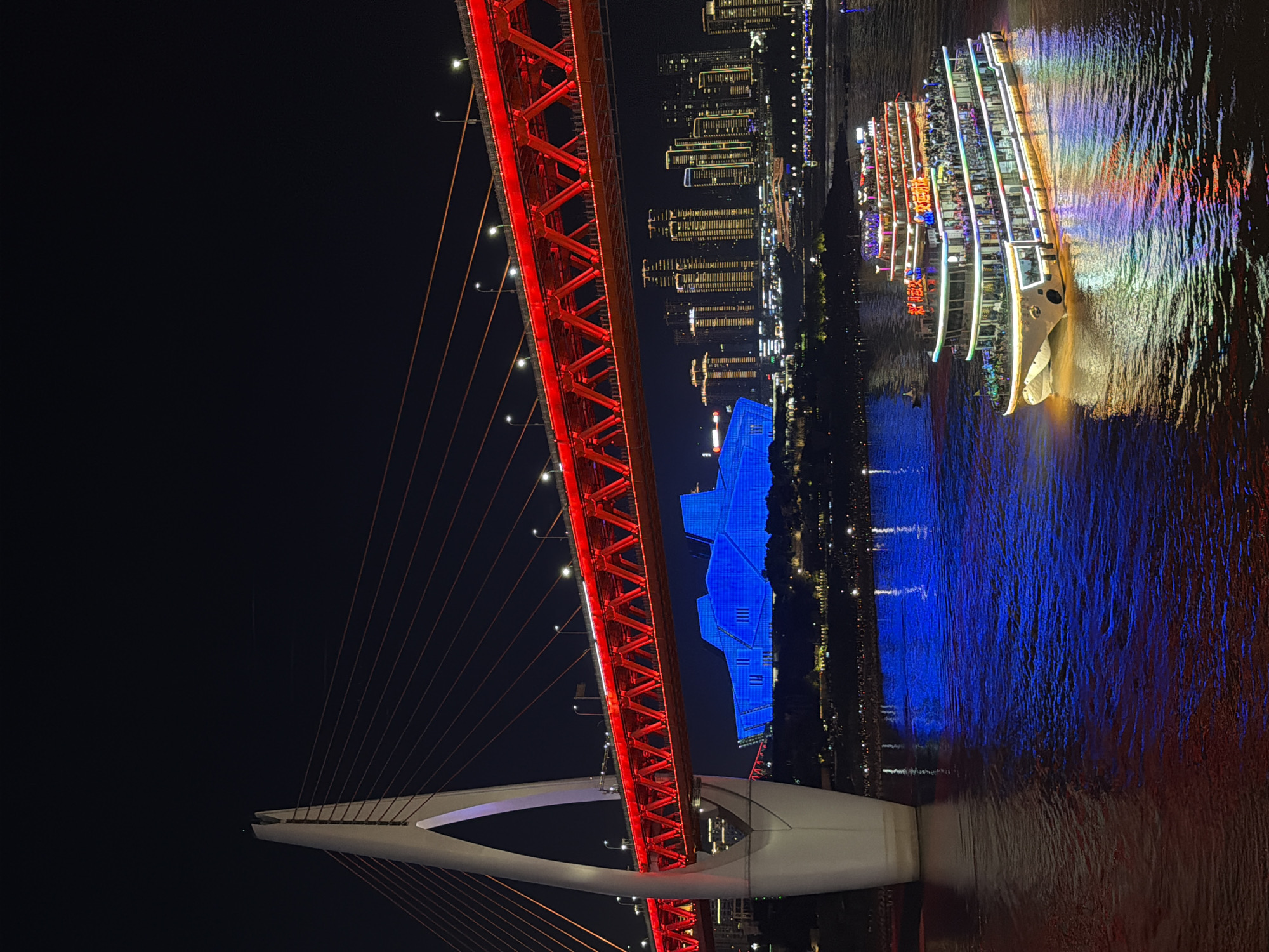 The image captures a nighttime scene of a city illuminated by vibrant lights. A prominent red suspension bridge, featuring a complex lattice structure and supporting cables, dominates the foreground. White supports of the bridge are also visible, arching into the dark sky.
To the right of the bridge, a large boat with multiple decks is illuminated by a colorful array of lights, casting reflections on the dark water. Passengers are visible on the decks of the boat. In the background, a cityscape of tall buildings with numerous windows lit up stands against the night sky. A striking blue-lit building, with a geometric design, is situated between the bridge and the cityscape, partially obscured by the bridge's structure. The water reflects the lights from the boat, buildings, and the bridge, creating a shimmering effect. The scene conveys a lively and festive atmosphere, suggesting a tourist attraction or a celebration. The time of day is clearly night, with a dark sky and artificial lighting being the primary light sources. The location is suggested to be Yuzhong District, China, implying an urban waterfront setting. Some Chinese characters are visible on the side of the boat, but they are not clearly legible.