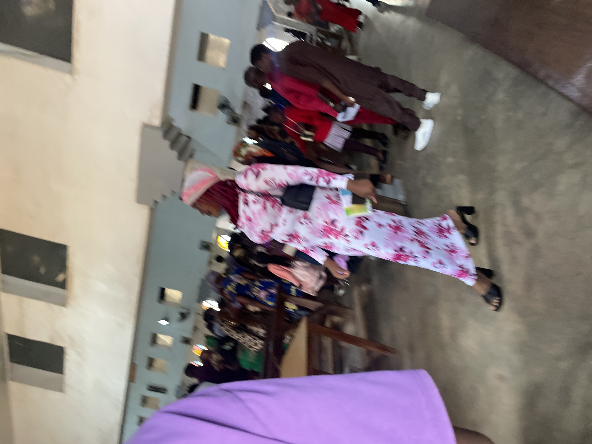 The image depicts an indoor gathering of people, likely in Jos, Nigeria. The environment appears to be a large hall with a concrete floor and high ceilings. Several windows are visible along the walls, some of which are dark. A staircase is also present in the background.
In the foreground, a person wearing a light purple garment is partially visible on the left. To their right, a woman in a white dress with a pink floral pattern and a matching pink and white headscarf is the primary subject. She is walking and carrying a black purse. She is wearing black heeled sandals.
Behind the main subject, a crowd of people is gathered, many of whom are wearing brightly colored clothing. Some individuals are standing, while others appear to be seated or moving. The overall impression is that of a community event or religious service. The lighting suggests it is daytime, possibly with artificial lighting supplementing natural light. The image is somewhat blurry, indicating motion or a lack of focus.