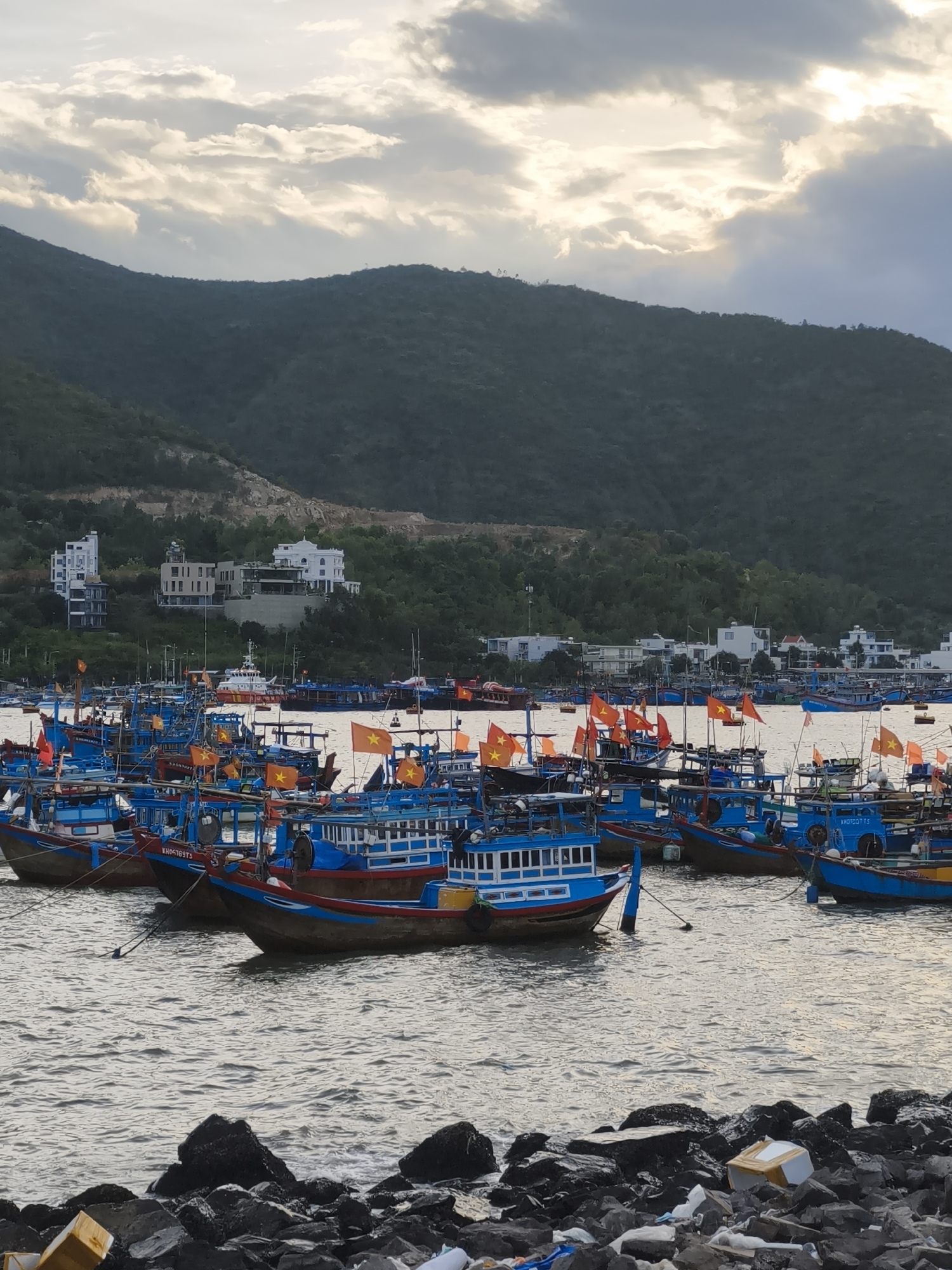 This image depicts a harbor filled with numerous small fishing boats, predominantly painted in vibrant blues and reds, docked together in the waters of Nha Trang, Vietnam. Many of the boats are adorned with Vietnamese flags, adding splashes of red and yellow to the scene. In the background, a lush green hillside rises, dotted with white buildings, suggesting a coastal city setting. The sky above is dramatic, with a mix of heavy, dark clouds and patches of bright sunlight breaking through, creating a somber yet striking atmosphere. The foreground shows a rocky shoreline with scattered debris, including plastic packaging. The overall impression is a bustling fishing port at dusk or during overcast weather.