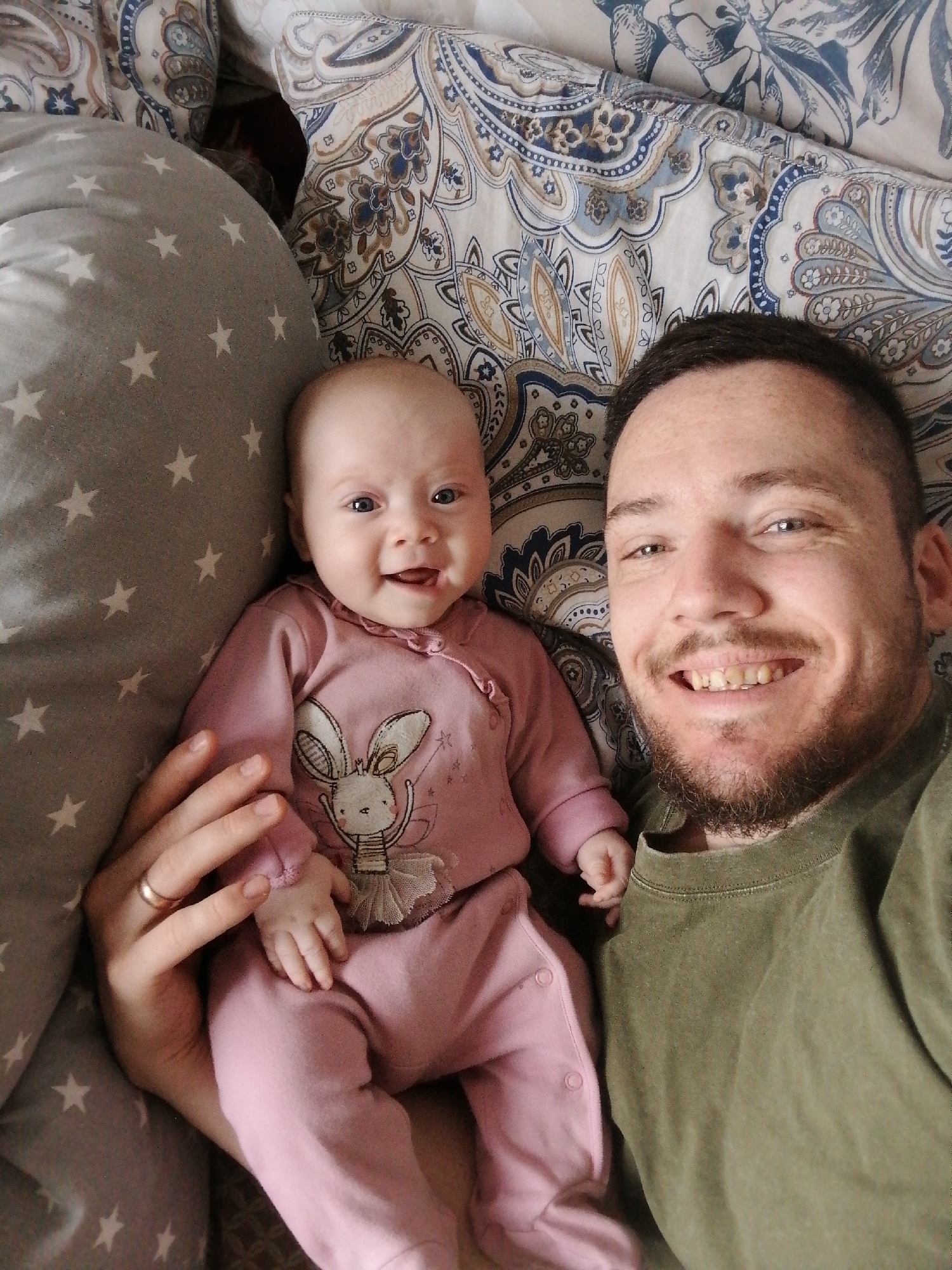 A close-up, top-down shot captures a man and a baby lying on a bed. The baby, dressed in pink pajamas with a bunny ballerina design, is positioned to the left of the man. The man is wearing a green t-shirt and has a beard. His hand is visible holding the baby. The background features bed linen with a gray star pattern and an ornate paisley design. The location is a private residence, city of Unknown, Unknown.