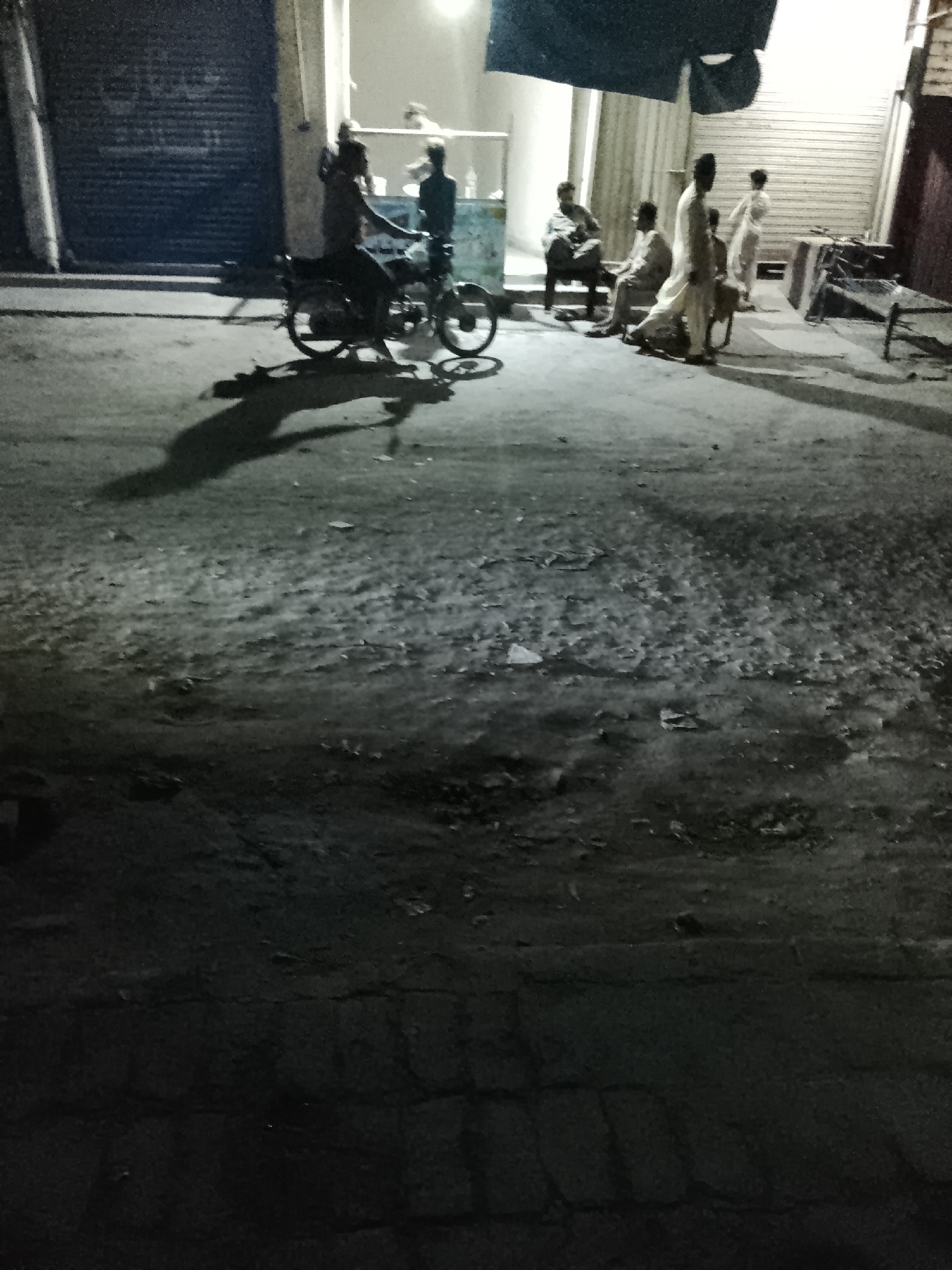This nighttime image depicts an informal gathering on a street or alleyway in Nasheman Colony, Pakistan. The scene is illuminated by a strong artificial light source emanating from an open doorway or shop in the background, casting long, stark shadows across the uneven ground.
In the mid-ground on the left, a dark-colored motorcycle is parked, with at least one individual, and possibly two, positioned on it. Their elongated shadows stretch towards the foreground. The ground itself appears to be a textured mix of dirt, debris, and worn-out pavement.
Towards the background, under the bright light, several men are gathered. Two individuals are seated on low chairs or stools, while another man, dressed in light-colored traditional attire, stands nearby. Additional figures are partially visible further back, suggesting a small community interaction or casual hangout.
The surroundings feature buildings with closed metal roll-down shutters, indicative of shops or commercial establishments. A dark fabric or awning hangs above the brightly lit area. The low light and resolution make details such as facial expressions and specific objects difficult to discern. Faint, illegible script, possibly in Urdu or Arabic, is visible on the shutter on the far left. The overall atmosphere suggests a quiet, social evening in a local setting.
