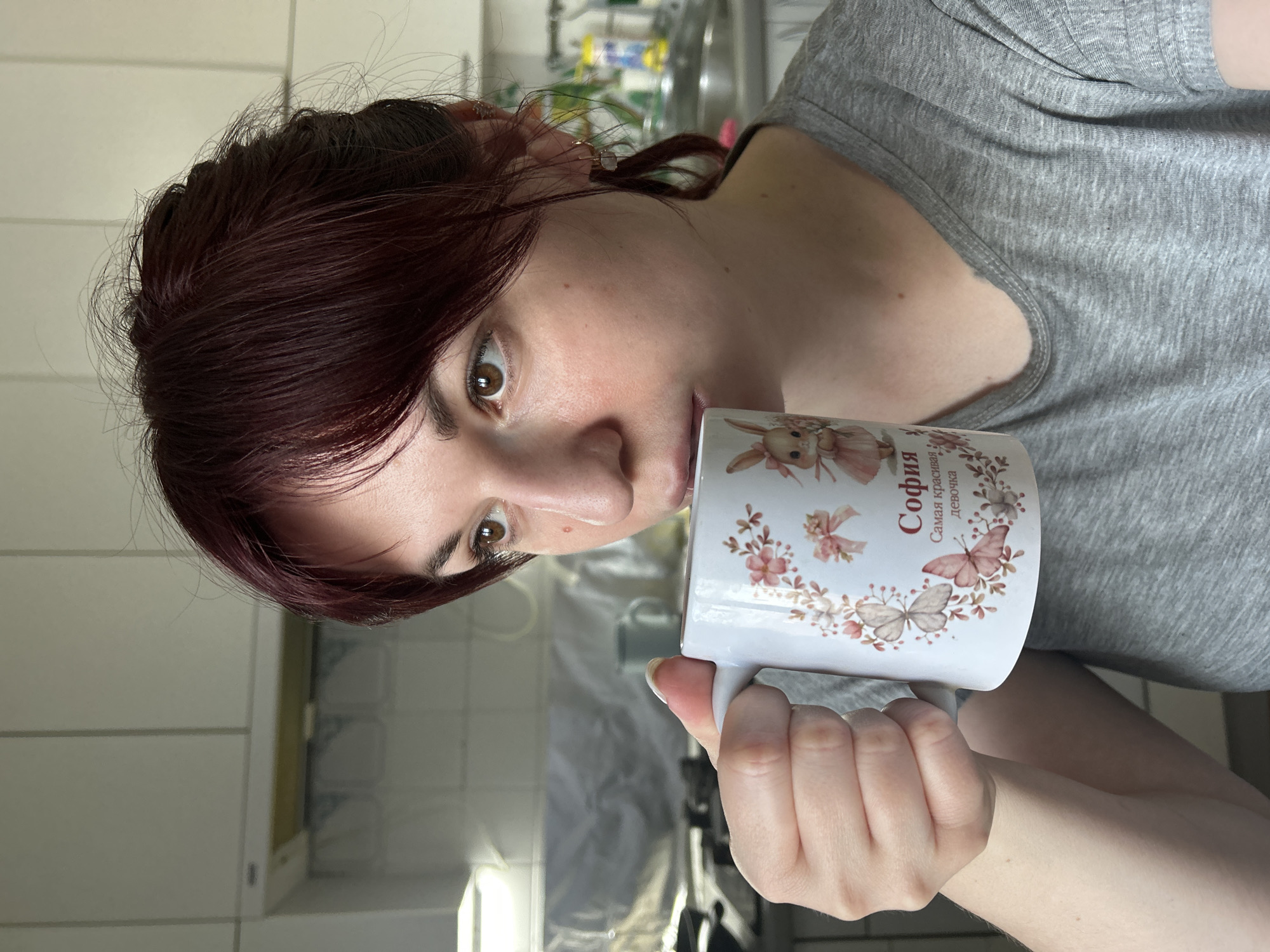 A woman with auburn hair drinks from a white mug with floral and butterfly designs and the name "Sophia" written in Cyrillic. She is wearing a grey t-shirt. The background shows a kitchen setting, possibly in a home, with cabinets and a sink visible. It is not possible to confirm the location in Фоки, Russia, based on visual data.