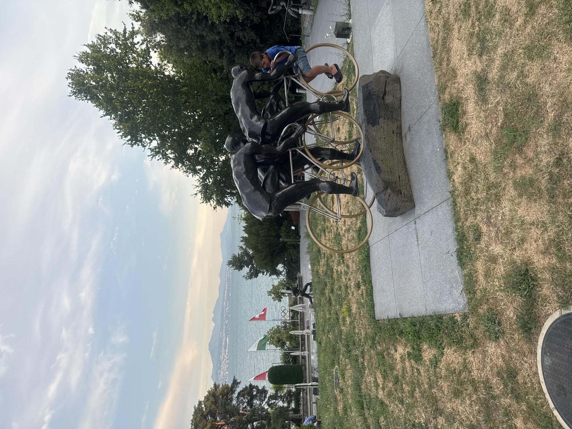 The image depicts a bronze sculpture of three cyclists in motion, situated in an outdoor setting by a large body of water, likely Lake Geneva, with mountains in the background. A young boy, wearing a blue t-shirt and shorts, is seated on the sculpture's handlebars, interacting with it. The sculpture rests on a stone base, which is surrounded by dry grass and a paved walkway. In the background, several flags are visible, including Swiss flags, and a statue of a runner. The sky is partly cloudy with a pale sunset glow. This scene is located at the Olympic Museum in Lausanne, Switzerland, a prominent city on the northern shore of Lake Geneva.