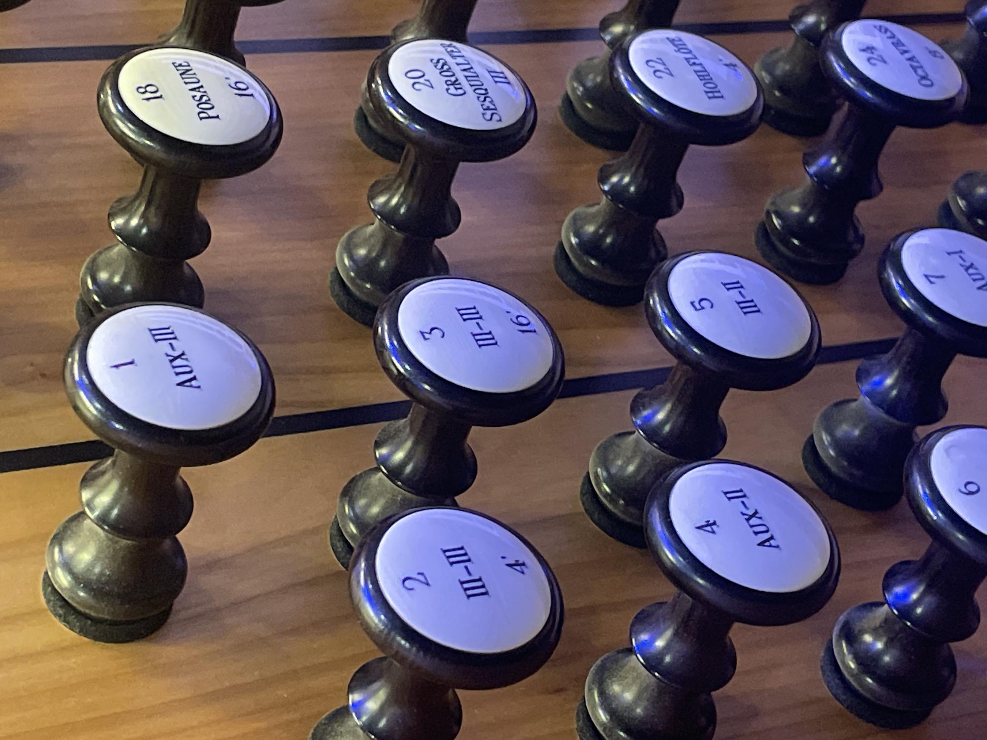 The image captures a detailed close-up of a section of a pipe organ console, specifically showcasing several dark wooden drawknobs, also known as stops, against a warm, light-colored wooden background. No people are visible in the frame. The scene is set in Yekaterinburg, Russia, focusing on the intricate controls of the musical instrument.
Each drawknob features a round, white face with black lettering, indicating its specific function, registration, or pipe rank. The lighting gives the white faces a subtle blue tint. The stops are arranged in rows, highlighting the complex array of sound options available to the organist.
Visible text on the drawknobs includes:
* "18 POSAUNE 16'"
* "20 GROSS SESQUIALTER III"
* "22 HOHLFLÖTE 4'"
* "24 OCTAVBAS 8'"
* "1 AUX-III"
* "3 III-III 16'"
* "5 III-II"
* "7 AUX-I"
* "2 III-III 4'"
* "4 AUX-II"
* A partial "6" is visible on a knob in the lower right corner.
The inscriptions denote various organ stops, with names like "Posaune" (a reed stop), "Hohlflöte" (a flute stop), "Octavbas" (an octave bass stop), and "Sesquialter" (a mixture stop). The numbers followed by an apostrophe (e.g., "16'", "4'", "8'") indicate the pipe length in feet, determining the pitch. Roman numerals often refer to the number of ranks in a mixture or specific organ divisions. "AUX-I," "AUX-II," and "AUX-III" likely represent auxiliary couplers or functions across different manual or pedal divisions. The image provides a clear view of the control interface of this large and complex musical instrument.