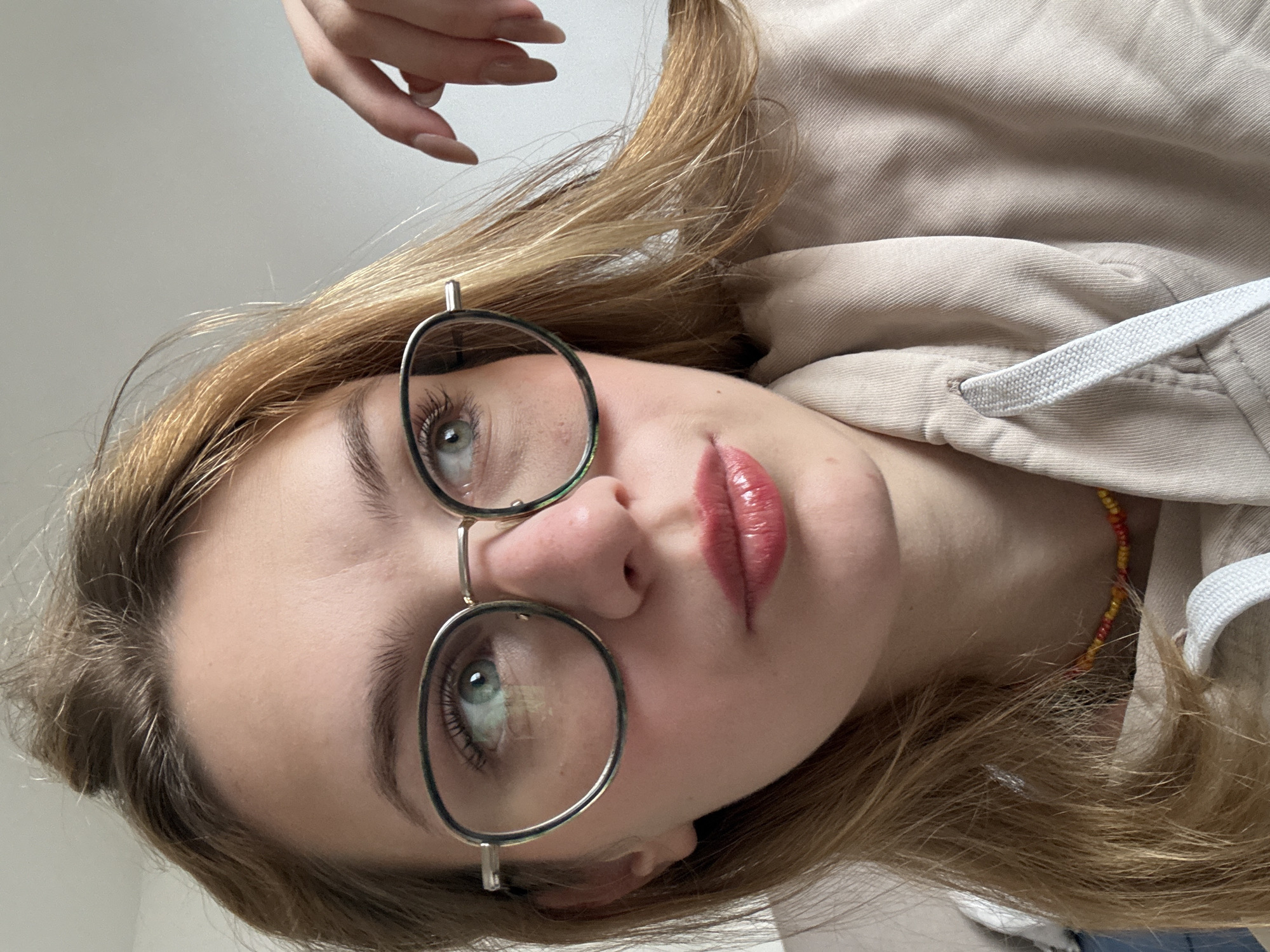 A young woman with fair skin and light brown hair is depicted in a selfie-style photograph. She is wearing round glasses with a silver frame. Her eyes are light green/blue. Her lips are a reddish-pink color. She is wearing a beige-colored top with a white drawstring. Her hand is visible at the top of the frame. There is a colored beaded necklace around her neck. The background is a plain white wall. The photograph is taken indoors, possibly in a private residence. There is no evidence to identify the location as Kragujevac, Serbia.