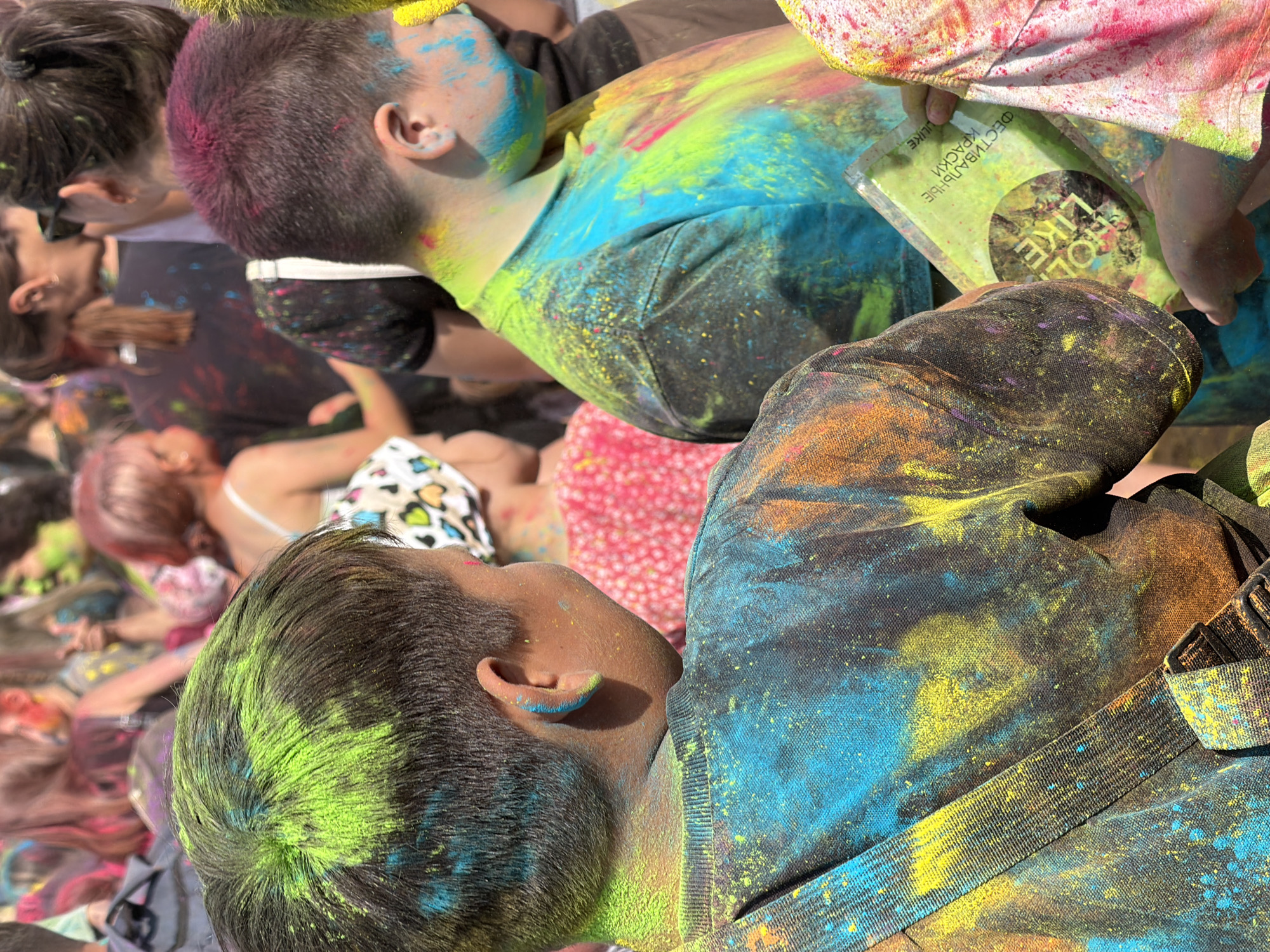 A group of young people are covered in brightly colored powder during a Holi festival celebration. The scene appears to be outdoors, with a crowd of people in the background. The main subjects are two boys in the foreground, both wearing dark t-shirts that are heavily dusted with powder in shades of green, yellow, blue, and orange. The boy on the left has green powder on his hair and a blue powder dusting his ear and neck. The boy on the right has pink and purple powder on his hair and his face and neck are smudged with blue and yellow powder. He is holding a small green package with Cyrillic text that appears to be a powdered dye packet. The lighting suggests it is daytime. The image was taken in Chaikovsky, Russia.
