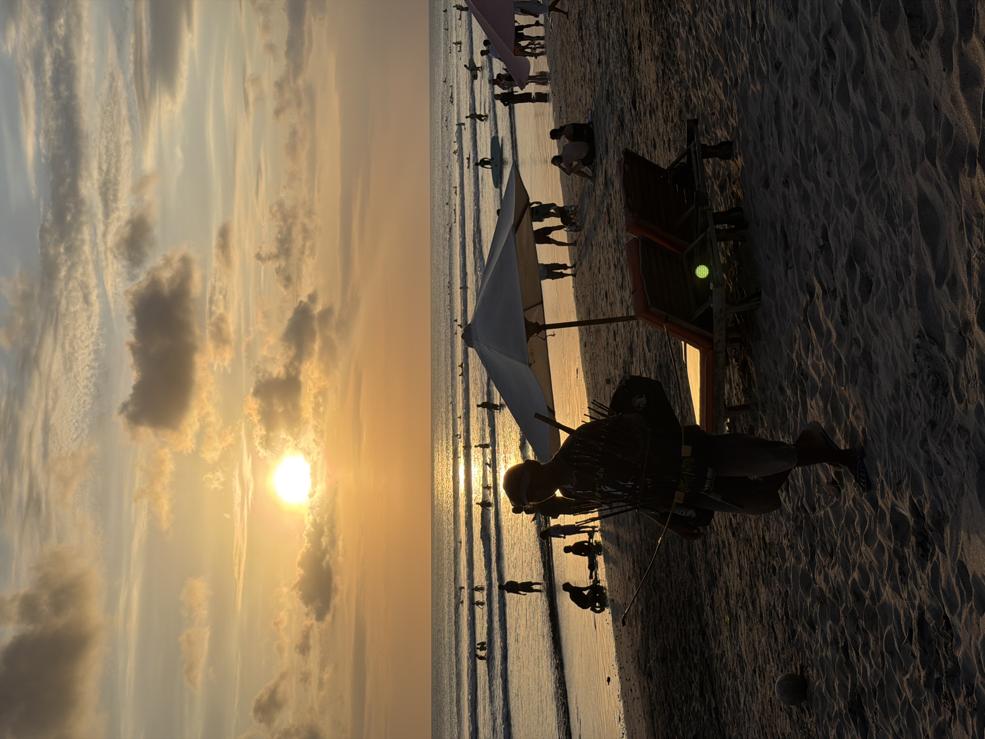 The image depicts a sunset on Kuta Beach, Indonesia. The sun, a bright orb, is setting on the left side of the frame, casting a warm, golden glow across the sky and reflecting on the ocean. The sky is filled with wispy clouds, illuminated by the setting sun in shades of orange, yellow, and soft gray.
The beach is populated by numerous people. Some are standing in the shallow water, while others are walking along the shore or sitting on the sand. Several individuals appear to be surfers or are holding surfboards, suggesting a surfing beach. In the foreground, a person is silhouetted against the sunset, wearing a hat and appearing to be holding fishing equipment. Adjacent to this person is a beach chair and an umbrella, common beach amenities. The sand shows the texture of footprints and waves, indicating it is a sandy beach.
The overall atmosphere is serene and relaxing, indicative of a pleasant evening at a popular tourist destination. The time of day is clearly sunset, and the weather appears to be clear, with the exception of the clouds. There is no visible text in the image. The scene is a typical depiction of a vibrant beach sunset with recreational activities taking place.