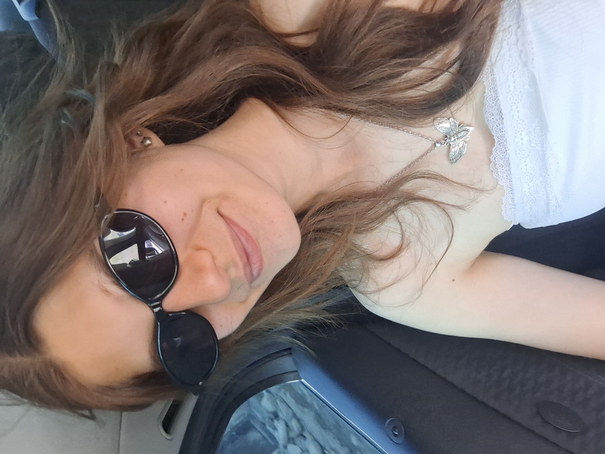 This media file is a close-up, horizontal, over-the-shoulder shot of a woman lying down, possibly in a car. The woman has long, brown hair that is partially covering her face. She is wearing sunglasses with black frames and a white top with lace detailing. A silver necklace with a butterfly pendant is visible around her neck. Her skin appears fair, and there are a few freckles on her cheek. The lighting suggests it is daytime. The background is dark and out of focus, but appears to be the interior of a vehicle, with a hint of rocky ground visible through what might be a window. There is no visible text in the image. The overall mood is casual and relaxed.