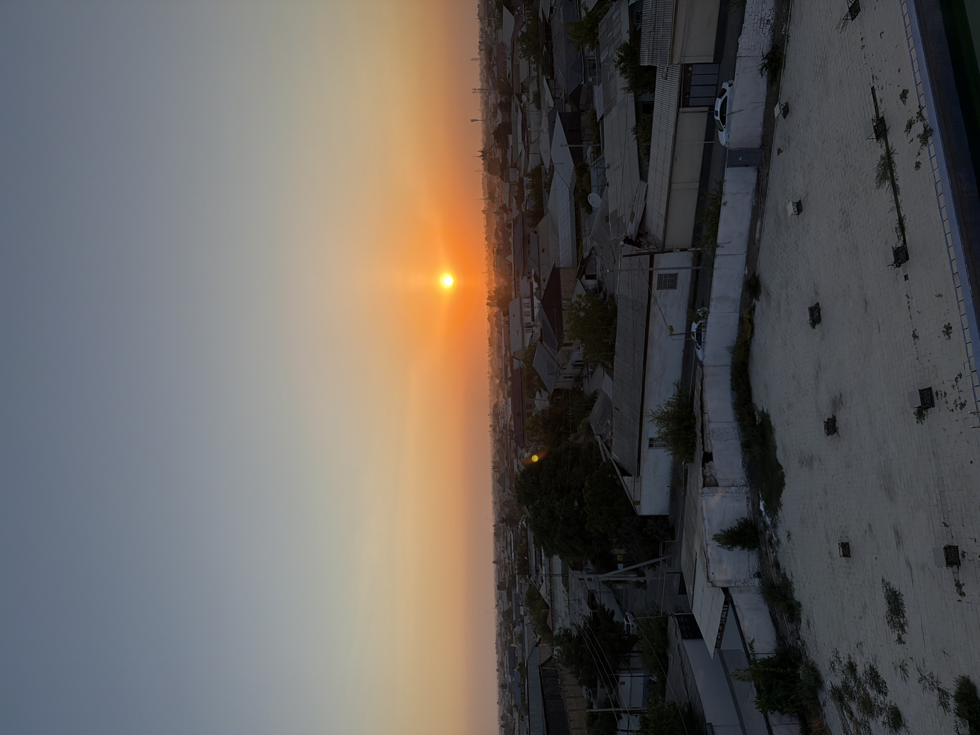 The image captures an aerial view of a cityscape at sunset in Andijan City, Uzbekistan. The sky is a gradient of pale blue at the top transitioning to a warm orange glow near the horizon, with the sun a bright, distinct orb near the center. The cityscape below consists of numerous low-rise buildings with predominantly gray, flat roofs, indicative of residential or commercial structures common in Central Asian cities. Some buildings are white-walled, while others have darker facades. Trees are scattered throughout the area, some appearing quite large and leafy, suggesting established neighborhoods. Roads and power lines are visible crisscrossing the urban landscape. In the foreground, there is a textured, light-colored rooftop with several square ventilation shafts or skylights. Two white cars are parked on a street in the mid-ground, visible between buildings. The overall impression is a calm, late-day scene in a moderately dense urban environment.