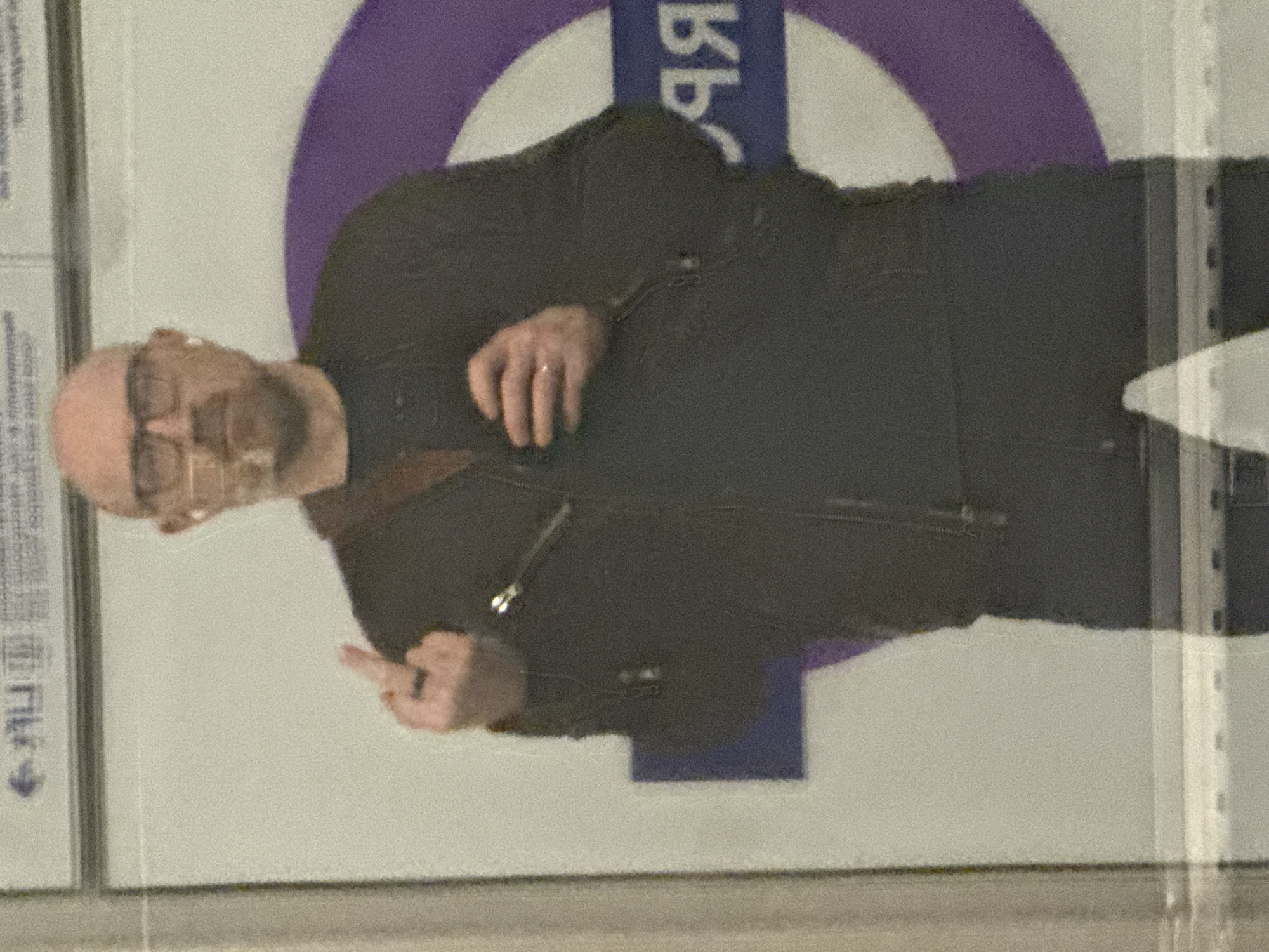 A man with glasses and a bald head, wearing a dark jacket, is visible through a pane of glass. He is giving the middle finger. Behind him is the London Underground logo, suggesting this might be occurring in London.
