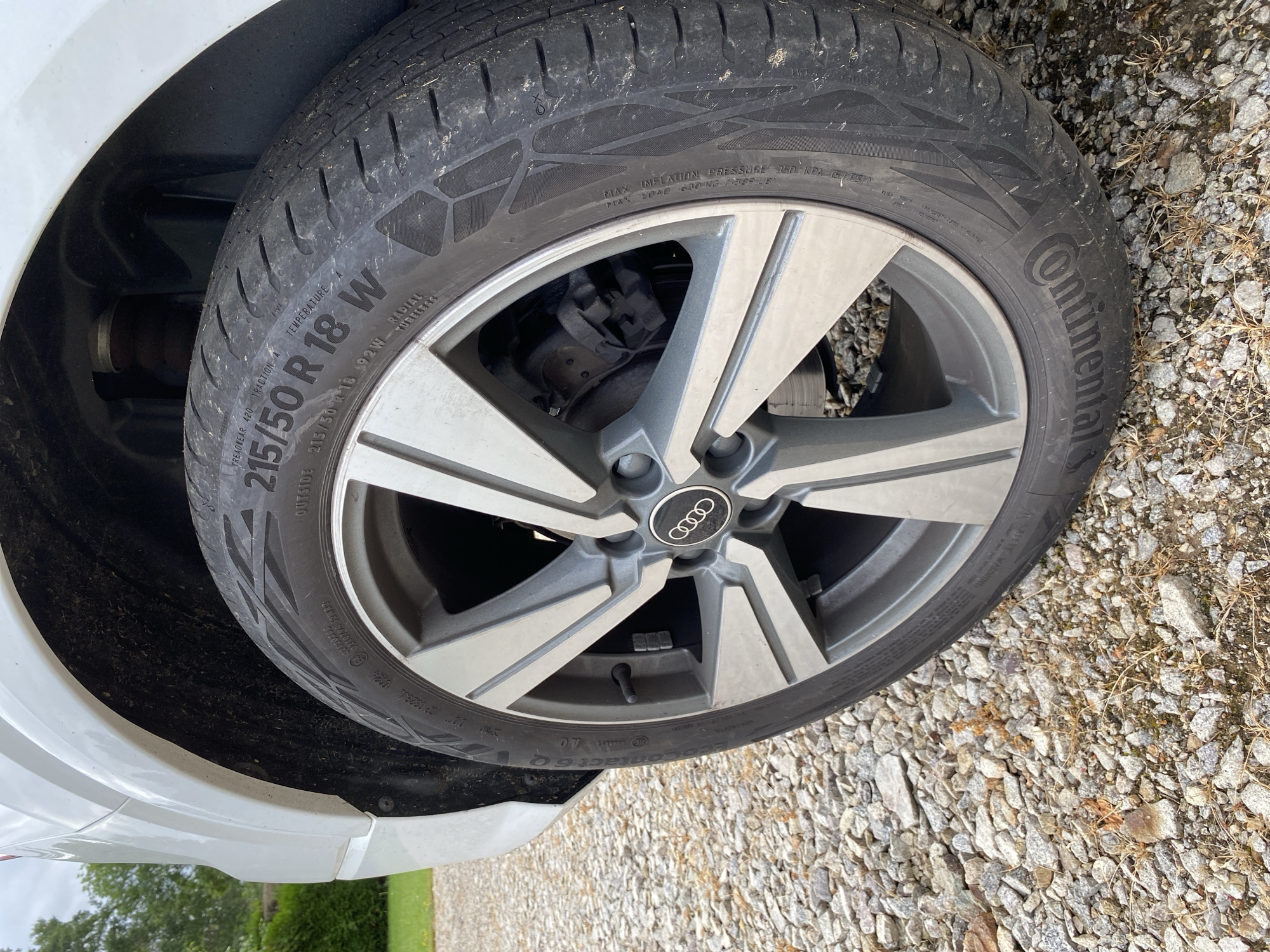 This image captures a close-up of the front wheel and tire of a white Audi vehicle, parked on a gravel surface. The scene is outdoors, brightly lit, suggesting daytime with fair weather. Green foliage and a hint of a tree are visible in the background, implying a setting in Courpière, France, possibly a driveway or rural road.
The primary subjects are the dark grey alloy wheel with the Audi rings logo prominently displayed on the center cap, and a black Continental tire. The tire tread pattern is visible and shows some wear and dirt. The tire also has a visible valve stem.
Notable details are the numerous markings on the tire sidewall, clearly readable from the image and its crops:
* Manufacturer: **Continental**
* Tire model: **ContiContact 6**
* Tire size specification: **215/50 R18 92W** (width 215mm, aspect ratio 50%, radial construction, 18-inch rim diameter, 92 load index, W speed rating).
* Tire performance ratings: **TREADWEAR 420 TRACTION A TEMPERATURE A**
* Maximum inflation pressure: **MAX INFLATION PRESSURE 350 KPA (51 PSI)**
* Maximum load capacity: **MAX LOAD 630 KG (1389 LB)**
* DOT code: **DOT JCP 0829JL 0824**, indicating the tire was manufactured in the 8th week of 2024.
* The word **"OUTSIDE"** is also visible, indicating the correct mounting direction.
No activity is taking place; the car appears to be stationary. The focus is purely on the technical details of the tire and wheel.