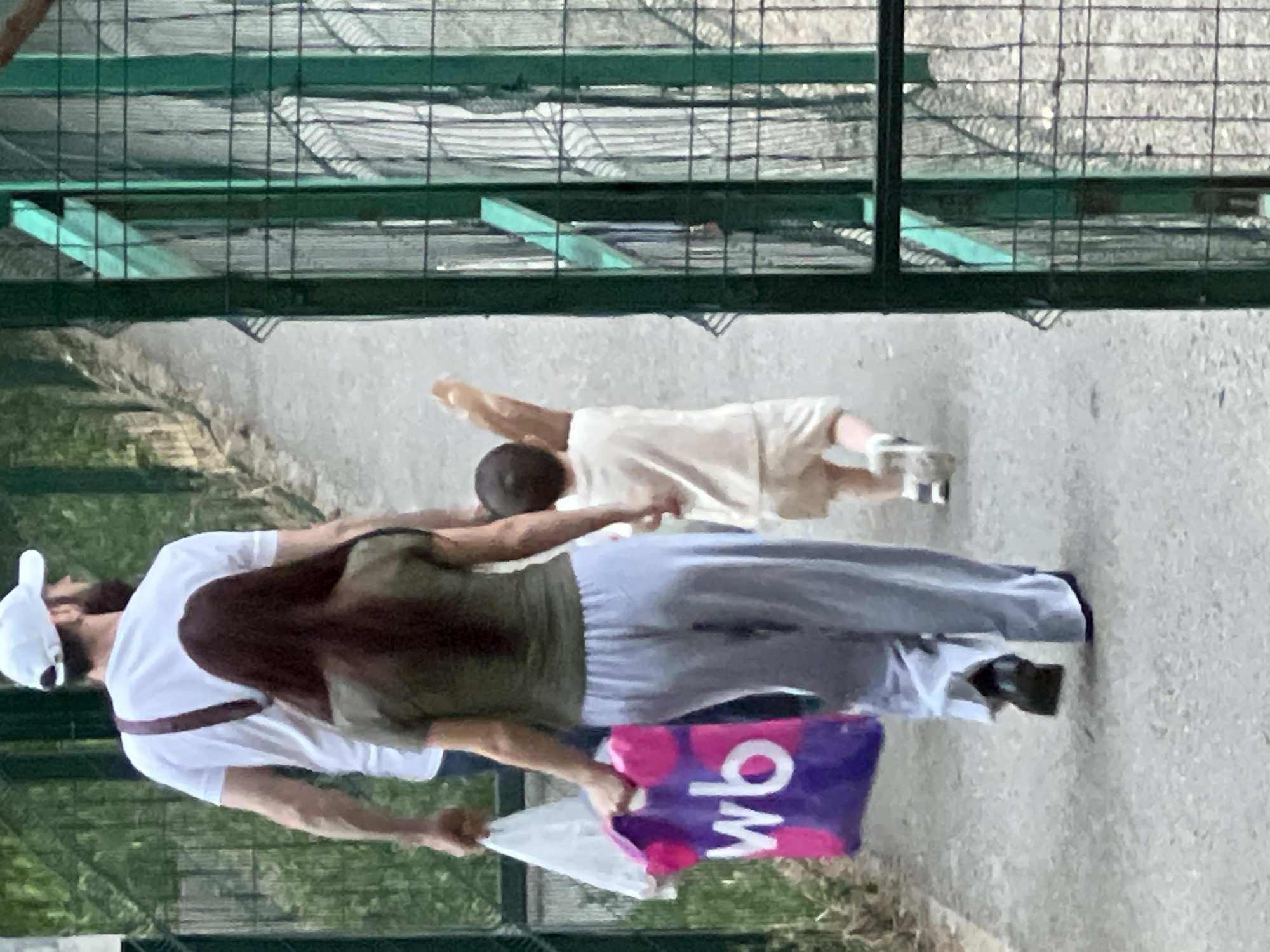 A family walks along a paved path outdoors. A man wearing a white t-shirt and a white baseball cap, with a beard, walks ahead. To his right, a woman in a green t-shirt and light blue pants carries a large purple shopping bag with pink polka dots and white lettering that says "wb". A young child in a cream-colored outfit walks ahead of the woman, holding a brown object that appears to be a baguette. The path is bordered by a green metal fence on the right, and foliage on the left. The setting appears to be a park or zoo in Krasnodar, Russia.
