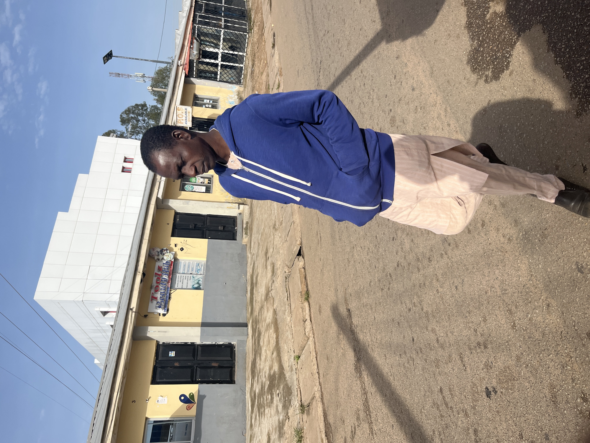 In the city of Jos, Nigeria, a man stands on a paved street. He is wearing a blue hooded sweatshirt and light-colored, striped trousers. The man is looking to his right and has his hands in his pockets. Behind him, there are several small buildings with yellow facades and dark doors. One building has a sign for "Toolz Beauty Hub." Further in the background, a larger, modern white building with geometric features is visible, along with a cell tower. The sky is clear and blue, with some scattered clouds. The ground is a mixture of asphalt and concrete, with some patches of dampness and scattered debris. Shadows from the man and other unseen objects are cast on the street.