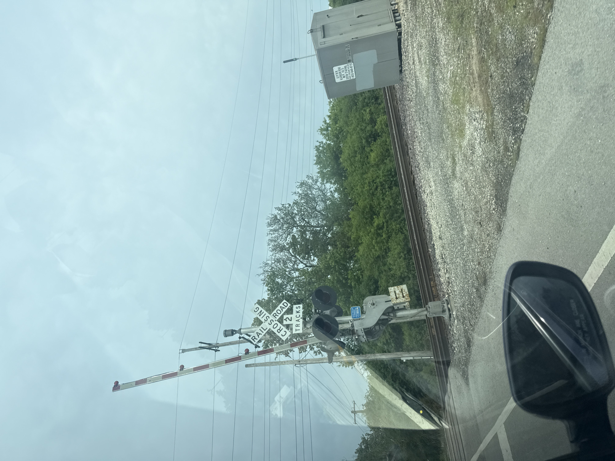 The image shows a railroad crossing in Des Plaines, Illinois, United States of America. The railroad crossing has a barrier arm, two signal lights, and a crossbuck sign indicating it is a two-track crossing. The crossing arm is lowered. The location is on or near Algonquin Road, at milepost 10.5. A gray rail transport container is beside the tracks. The image was taken from a vehicle. The vehicle's side mirror is visible in the bottom right of the frame. Overcast sky. Trees line the railway.