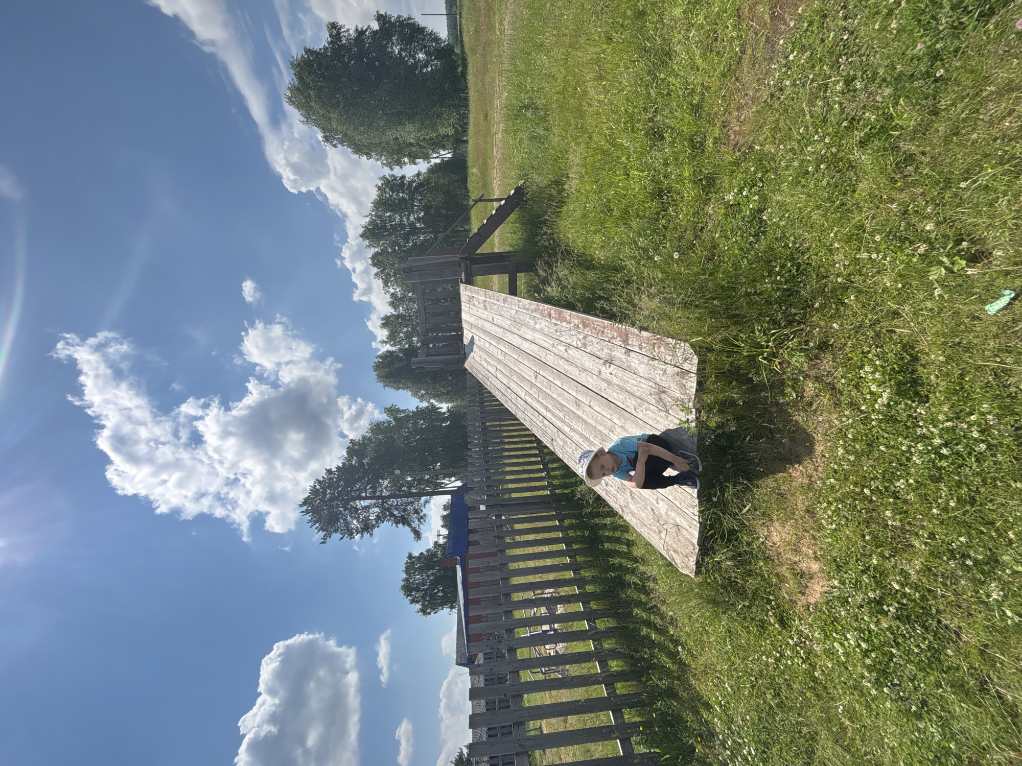 A young boy wearing a light blue t-shirt, dark pants, a light-colored hat, and dark shoes sits on the end of a wooden slide in a grassy outdoor area. The slide is constructed from light-colored wooden planks and has stairs leading up to it on the right side. The boy is facing away from the camera, with his knees tucked up towards his chest, and his arms wrapped around his legs. He appears to be looking downwards.
Behind the slide and to the left is a wooden fence with vertical posts and horizontal planks, and behind the fence, a red building with a blue roof is visible. The environment is rural, with a large expanse of green grass in the foreground and middle ground, dotted with small white flowers. In the background, numerous trees, both deciduous and coniferous, are present. The sky is bright blue with scattered white, fluffy clouds.
The image depicts a scene in a village or rural settlement, consistent with the landscape typically found in the outskirts of a city like Kurgan, Russia. The overall atmosphere is one of a sunny, summer day. There are no other people visible in the image. The lighting suggests it is daytime, likely late morning or early afternoon, with the sun casting shadows from the slide onto the grass.