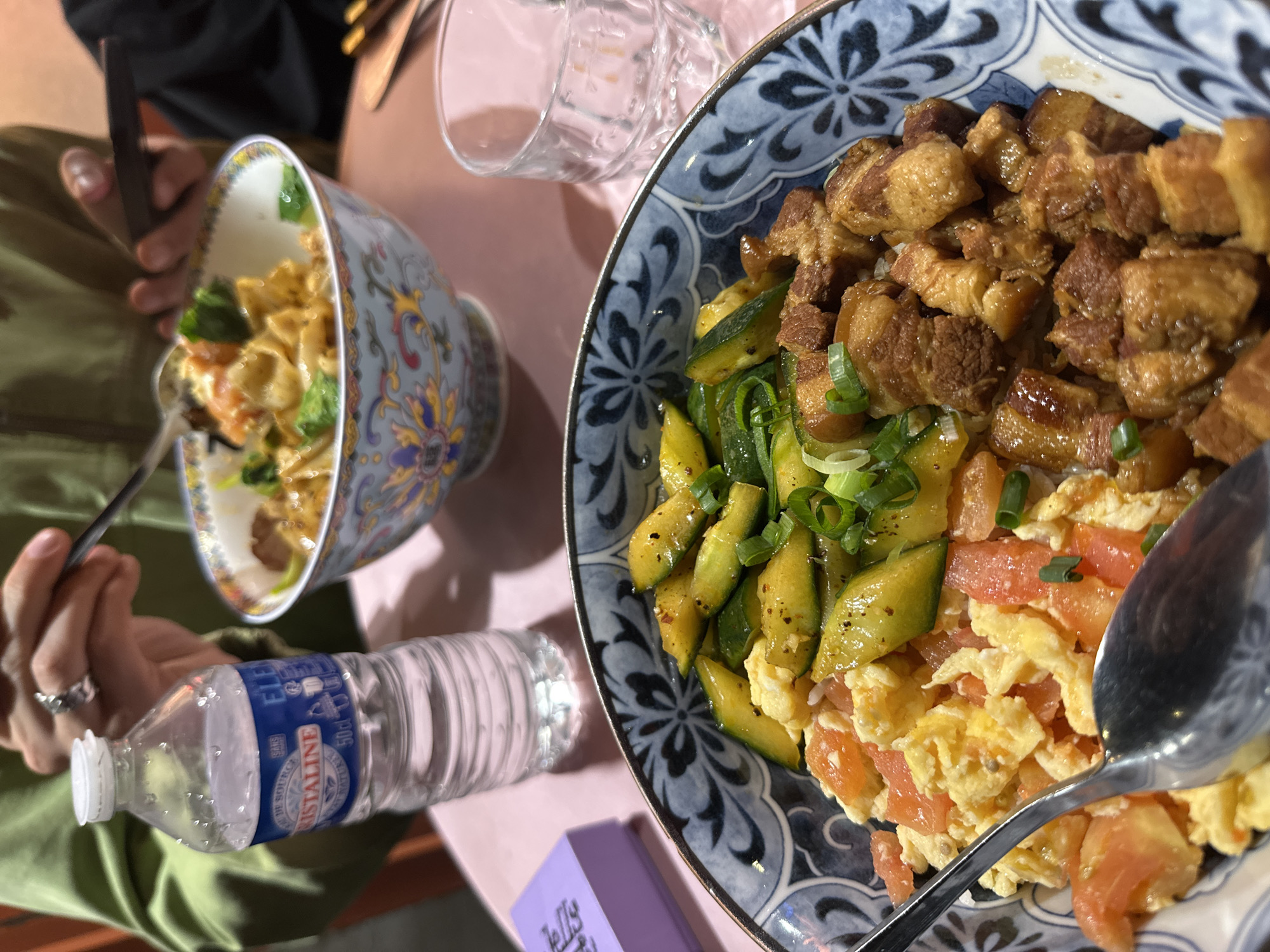 Two individuals are seated at a light pink table, engaged in a meal. The scene captures a top-down view, focusing on the food and hands.
In the foreground and to the right, a large, ornate blue and white patterned bowl holds a substantial meal. This bowl contains a portion of white rice topped with generous cubes of braised pork belly, glistening with sauce. Alongside the pork, there are slices of seasoned cucumber and a mix of scrambled eggs with diced tomatoes, garnished with chopped green onions. A silver spoon rests within this bowl, ready for eating.
To the left, a second person holds a smaller, elaborately decorated bowl with a yellow, blue, and red floral pattern. This bowl appears to contain noodles with green vegetables like broccoli or similar greens, and possibly some protein or tofu. The person is holding a dark utensil, likely chopsticks, and a dark-colored smartphone. Their left hand wears a ring.
Between the two diners, a transparent plastic bottle of water is visible. The label clearly reads "CRISTALINE," "EAU DE SOURCE," and "50cl," along with "SANS GAZ." A clear glass is also on the table, near the blue patterned bowl. A small, partially visible purple box with the word "Jelly..." on it is situated near the blue bowl.
The lighting is warm and artificial, suggesting an indoor setting, possibly a restaurant or home. The brand of water bottle is French, aligning with the provided context of Paris, France. The overall impression is a casual dining experience.