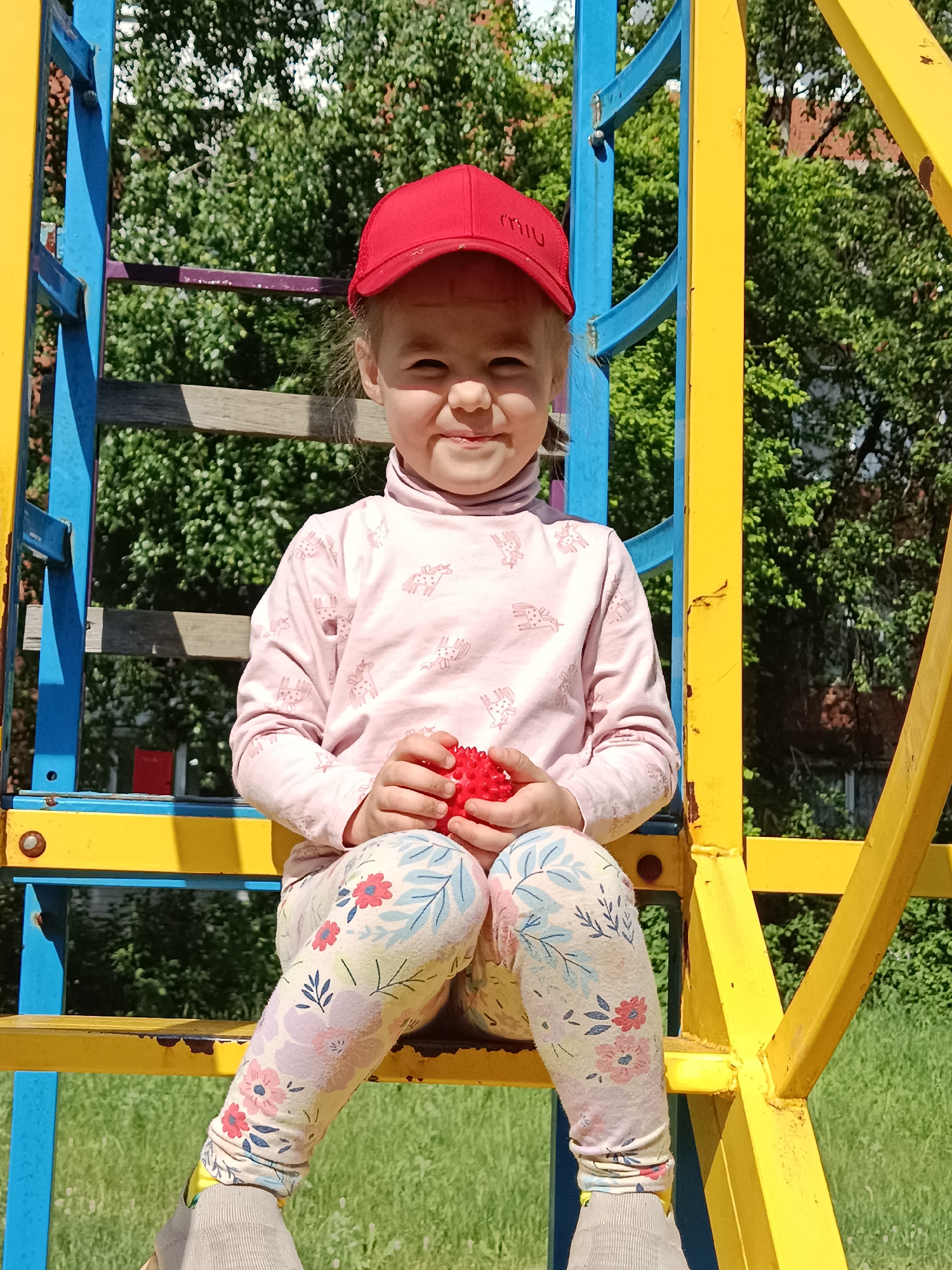 A young girl is depicted sitting on a colorful playground structure outdoors on a sunny day. She is looking directly at the camera with a slight smile or smirk. The girl is wearing a red baseball cap with white text that appears to read "miu" on the front. Her long-sleeved top is light pink with repeating light brown or beige animal (possibly deer) patterns. She wears floral-patterned leggings with various shades of pink, blue, and green on a light background. Her feet are visible, covered by light grey socks or shoes.
In her hands, she holds a small, round, red spiky ball. The playground structure is made of metal bars, painted in alternating bright yellow and blue, with some wooden planks visible behind her. The background consists of lush green foliage from trees and bushes, indicating a park or natural setting. Green grass is also visible below the structure. The bright sunlight suggests it is daytime. The overall impression is of a child enjoying herself in a playful environment.