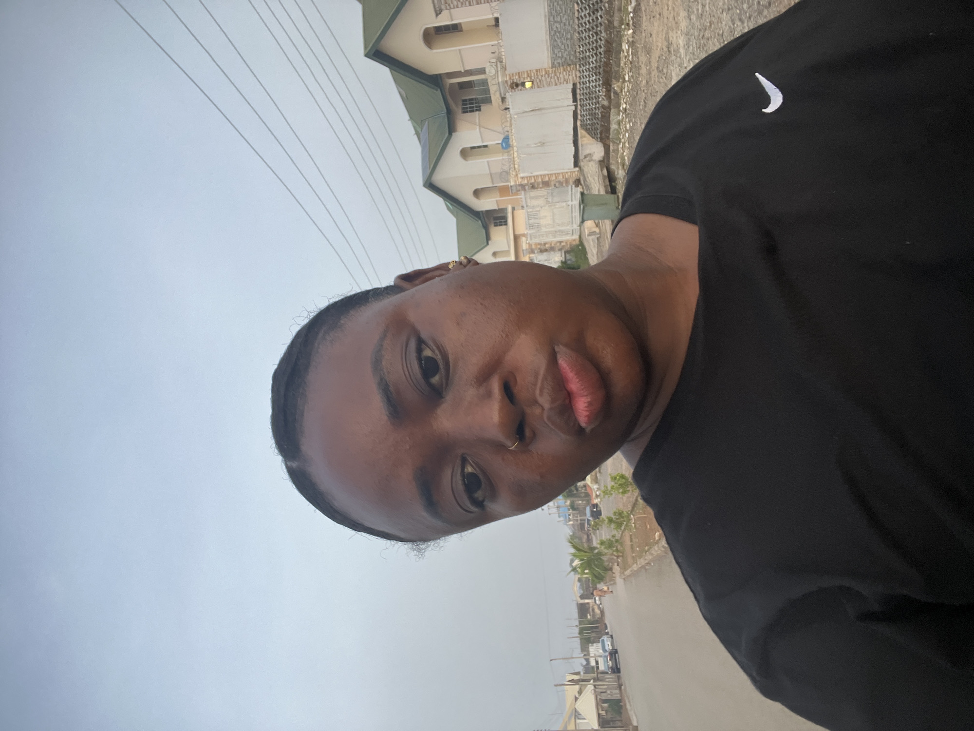 A person is shown in a selfie. The person has brown skin, dark hair pulled back, and is wearing a black t-shirt with a white Nike logo. They have a nose ring. In the background is a street in Abuja, Nigeria. There are buildings, cars, and telephone poles visible. The sky is overcast.