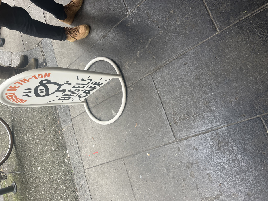 An overhead shot captures a street-level scene on a dark gray tiled pavement in Genève, Switzerland. In the upper left, two sets of human legs are visible. One person wears black trousers and light brown lace-up boots. Another pair of dark boots is partially visible further left, suggesting a second person.
The central focus is a white, oval-shaped A-frame sidewalk sign with a rounded white metal base. The sign's graphic features a stylized, black-outlined monkey-like creature with a large, smiling mouth. Above the creature, red French text reads "OUVERT DE 7H-15H," indicating opening hours from 7 AM to 3 PM. Below the creature, vertically aligned, are the words "BAGELS CAFE" in black, suggesting the type of business.
To the far left, a portion of a bicycle wheel and its frame are visible. The ground consists of large, rectangular dark gray pavers with visible grout lines. Some areas of the pavement appear damp or discolored, possibly indicating recent moisture. The overall setting is an urban sidewalk, likely in front of the Bagels Cafe advertised by the sign. The image suggests a daytime scene, though the exact lighting conditions are not strongly indicative of a specific time. No specific emotions are discernible.