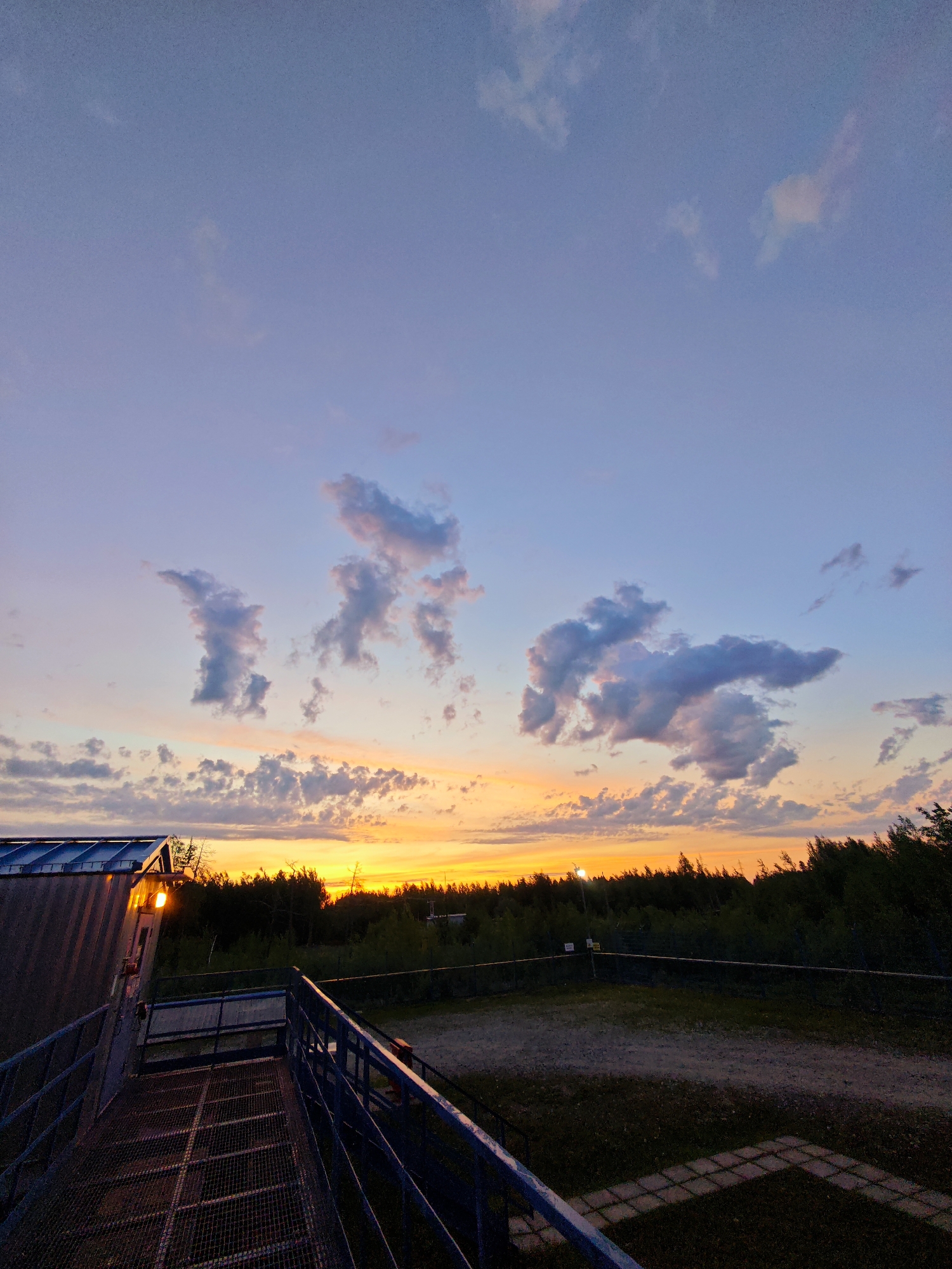 The image depicts a sunset over a treeline, with a metal structure in the foreground. The sky is a gradient of blue and pink, with scattered clouds. A building with a slanted roof is visible on the left, and a metal walkway with railings extends across the foreground. The treeline in the distance is dark, silhouetted against the bright orange and yellow hues of the setting sun. The ground in front of the treeline appears to be a dirt path, with some patches of grass and paving stones. The overall scene is peaceful and serene, with a sense of natural beauty.