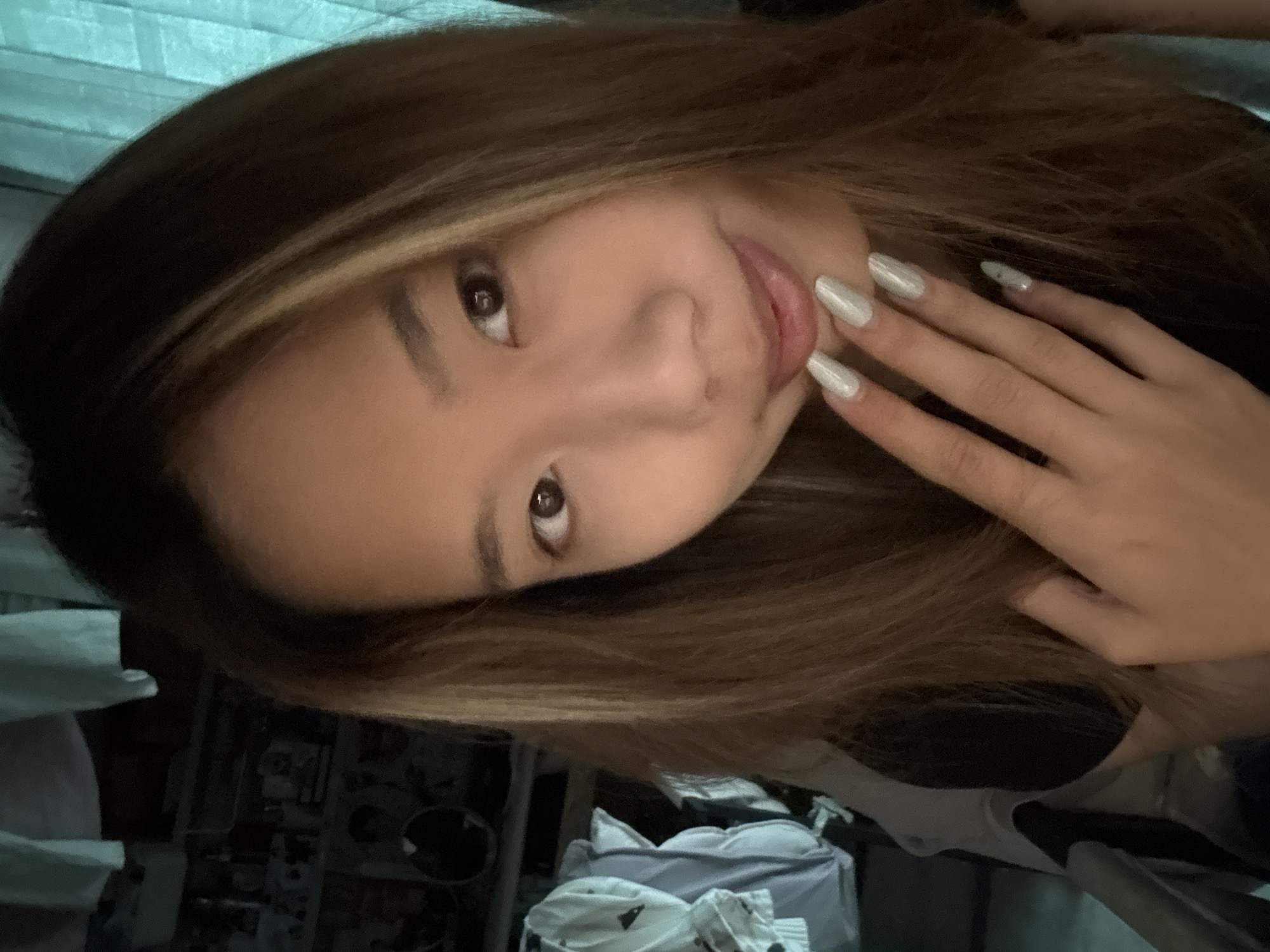 A medium shot selfie features a young woman indoors, her face filling the majority of the frame. Her long, light brown hair with blonde highlights is parted to the side and frames her face. She has brown eyes and is looking towards the camera with a slight smile. Her right hand, with neatly manicured pale green nails, is raised to her chin. In the background, out of focus, are various items including clothing hanging on a rack, shelves with what appear to be framed pictures or posters, and a teal-colored curtain. The lighting suggests an indoor environment, possibly a bedroom or closet. There is no discernible information within the image that would place the location in Nan'an District, China.