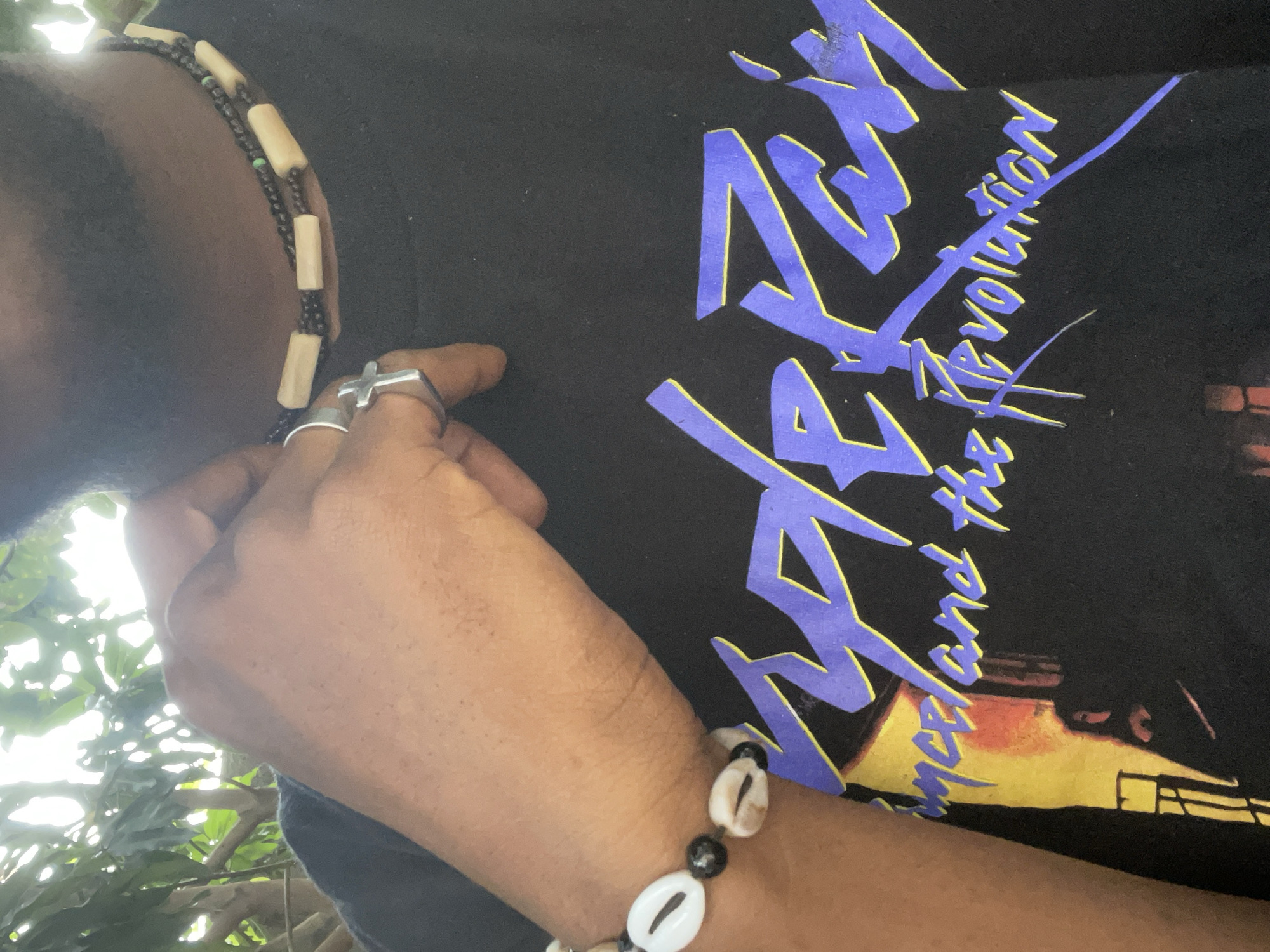 A medium shot depicts a person wearing a black t-shirt with a blue and yellow graphic design, a beaded necklace, a ring, and a beaded bracelet. The shirt reads "Prince and the Revolution." The person's left hand is raised, with fingers touching the shirt. The necklace consists of small black beads interspersed with larger, light-colored cylindrical beads. The bracelet features white shells and black beads. The setting appears to be outdoors, with greenery visible in the background. The location is not determinable based on the image.