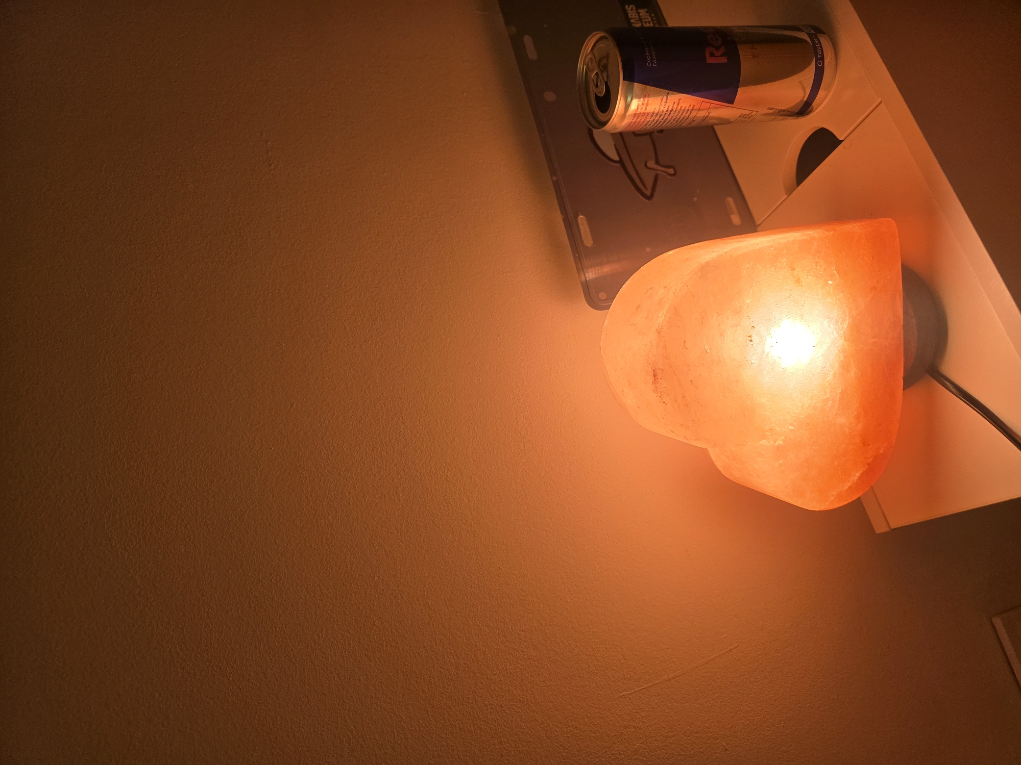 A heart-shaped Himalayan salt lamp, glowing with an orange hue, sits on a white surface. Beside it, a Red Bull energy drink can is positioned. In the background, a metal license plate featuring a UFO graphic is leaned against the wall. The wall itself is a textured, warm-toned color. The scene suggests an indoor setting, possibly a bedroom or a living area, with the dim lighting creating a cozy atmosphere. There is no indication of Golyanovtsi, Bulgaria, in the image.