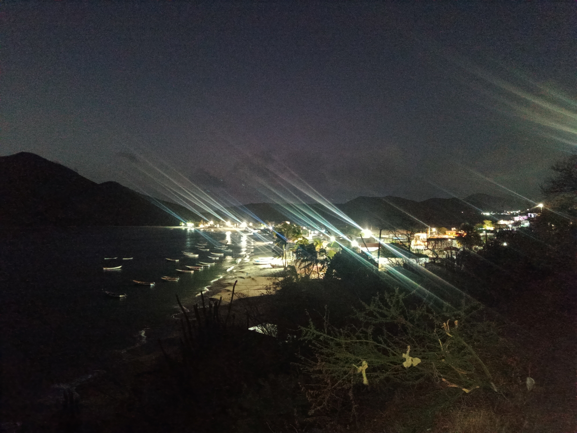 A nighttime image shows a coastal scene in Juan Griego, Venezuela. The perspective is from an elevated position, looking down at a bay with a shoreline. Several small boats are moored in the water near the beach. The shoreline is lit by streetlights and buildings of a town or village visible along the coast. The lights create prominent beams that extend upwards into the night sky. Hills or low mountains are silhouetted against the dark sky in the background. Bushes and vegetation are in the foreground.
