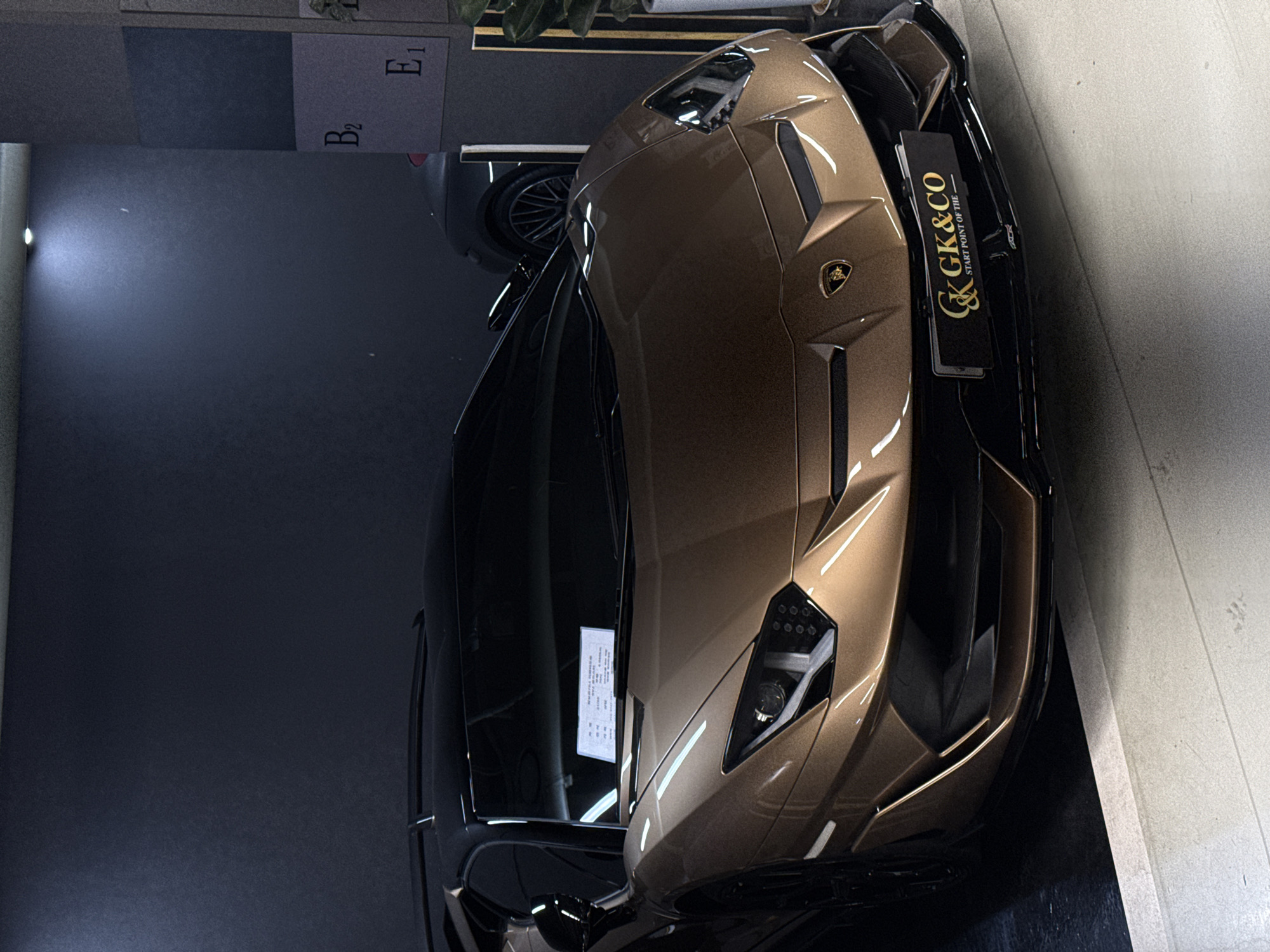 A high-angle indoor shot shows the front portion of a bronze-colored Lamborghini sports car parked inside what appears to be a showroom or garage, possibly in Seoul, South Korea. The car is angled towards the right with the front bumper near a white wall. The left side of the frame features a dark background, and the upper-left corner shows a section of a wall with what looks like color swatches or labels. The car has a "GK&CO" placard on its front, indicating a dealership or automotive company. A registration or sales document is seen adhered to the windshield on the driver's side. A part of another car is visible in the background.