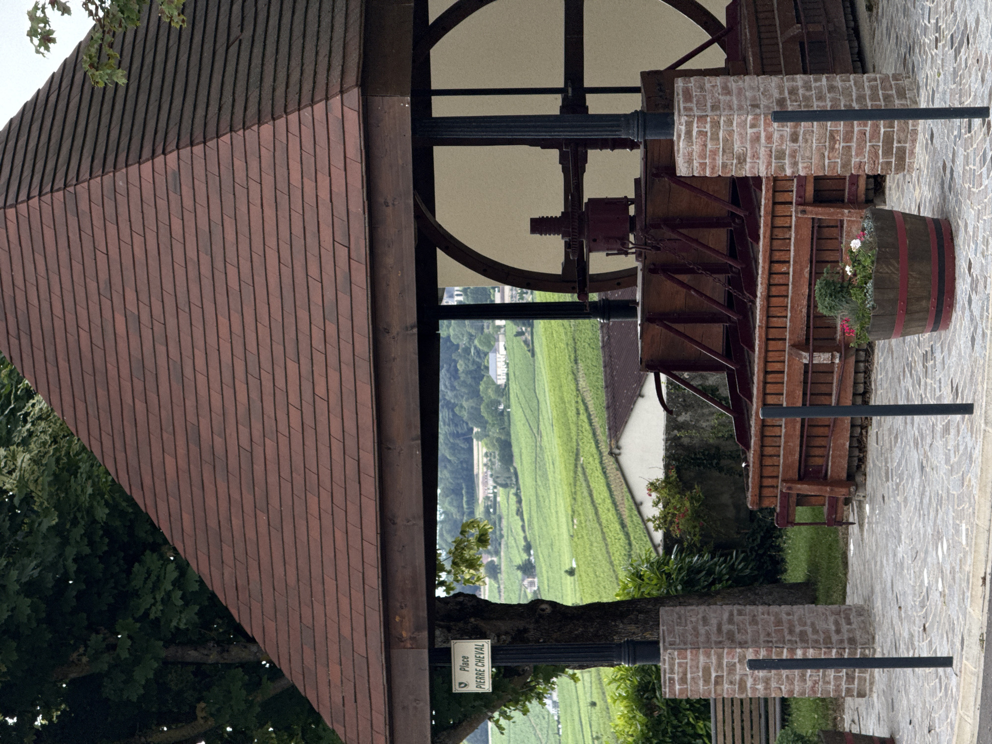 This image captures a static, serene outdoor scene featuring a historical winemaking exhibit set against a vast vineyard landscape. In the foreground, a substantial open-air structure is visible, characterized by a sloping roof made of reddish-brown tiles. Underneath this roof, an impressive, antique mechanical wine press dominates the space. This press is constructed from robust wooden beams, features a large circular wooden frame, and incorporates intricate metal components, including gears and shafts, suggesting its historical function in grape processing.
To the right of the press, a decorative wooden barrel, adorned with red and black stripes, serves as a planter, blooming with white and pink flowers and lush green foliage. The ground around the structure is paved with light-colored cobblestones. Several brick pillars, built from light red bricks, and black metal posts are also present, adding to the structural elements of the scene.
Beyond the foreground structure, a breathtaking vista of vibrant green vineyards unfolds across rolling hills, extending deep into the background. Scattered white buildings and houses are nestled amidst the grapevines, highlighting a settled agricultural landscape. The sky above appears bright, possibly overcast, providing soft, diffused light.
A prominent detail is a white rectangular sign attached to a black post, bearing the text "Place PIERRE CHEVAL". A large tree with green foliage is also visible, partially obscuring the view of the vineyards. The overall setting strongly evokes a sense of history and tradition associated with viticulture, consistent with the provided location context of Champillon, France. No people or activities are depicted, giving the scene a timeless and tranquil atmosphere.