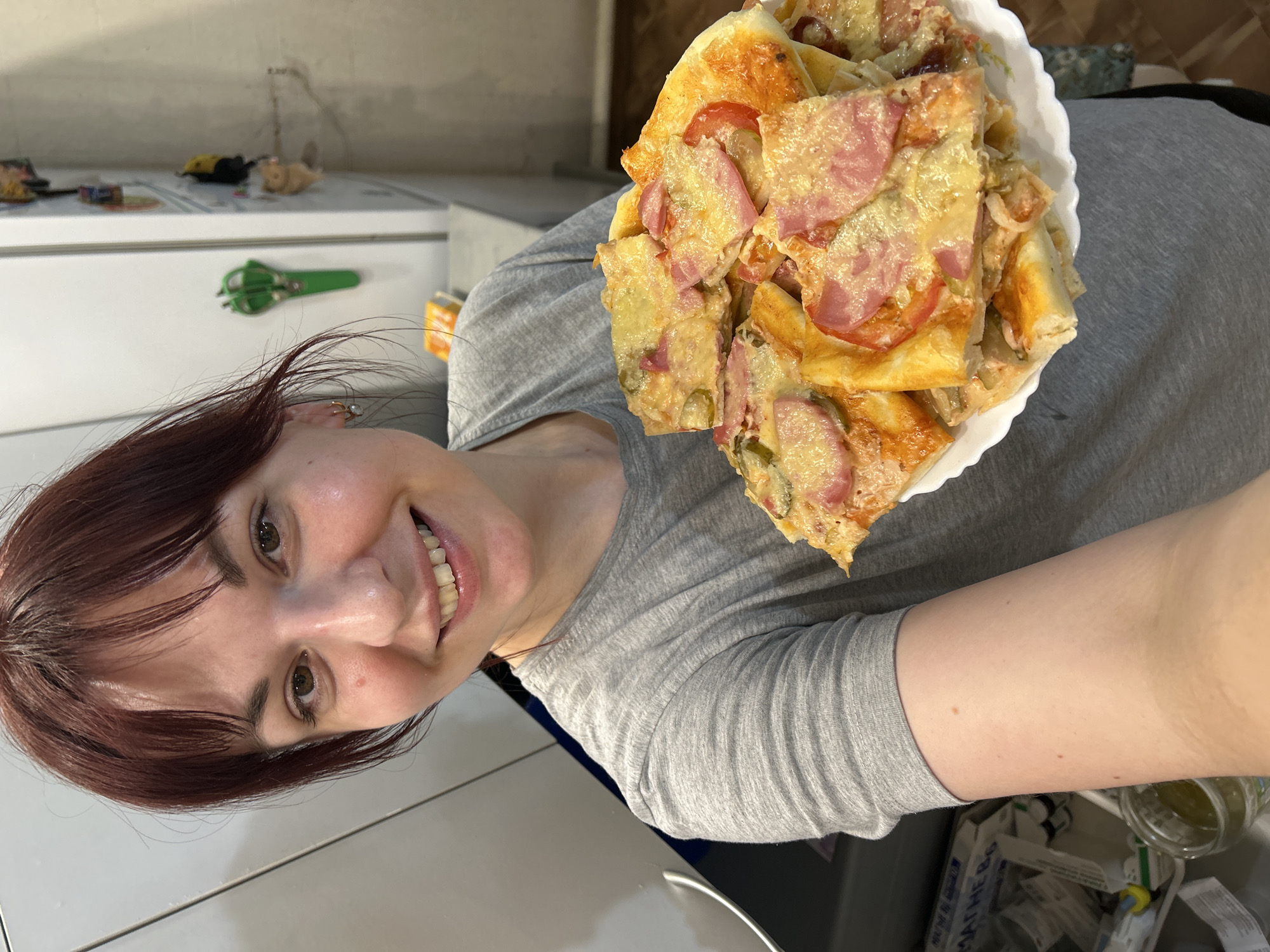A woman with auburn hair, brown eyes, and a wide smile holds a white plate piled high with what appears to be homemade pizza. The pizza is cut into irregular square pieces, topped with melted cheese, ham, and tomatoes. The woman is wearing a gray, long-sleeved shirt. The background includes a white kitchen counter with objects such as scissors and toys, and part of a wall. Based on the location information provided, the scene is assumed to be in Фоки, Russia.
