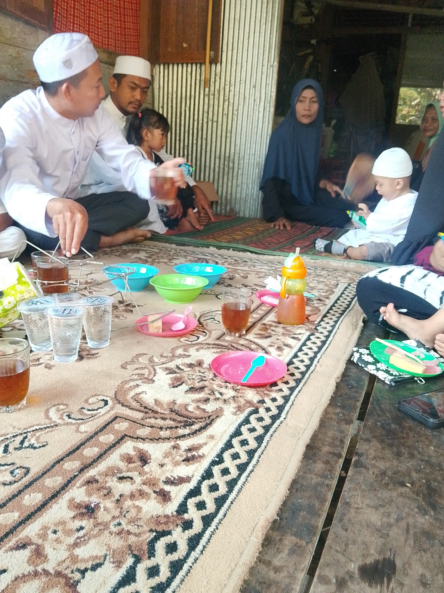 This image depicts a gathering of people, likely a family, seated on a rug inside what appears to be a modest dwelling in Buluan, Indonesia. The room has a rustic aesthetic, with a corrugated metal wall and wooden elements visible.
In the foreground, on the rug, are several items laid out for what seems to be a meal or snack. There are clear plastic cups filled with a dark liquid, likely tea, and smaller plastic bowls in blue and green. A pink plate holds a small slice of cake and a pink spoon. Another pink plate with a turquoise spoon is visible further to the right. A bottle with a yellow and orange dispenser top, filled with a brown liquid, is also present.
Several individuals are visible in the scene. A man wearing a white cap and a white long-sleeved shirt is seated on the left, appearing to pour something from a small container into a glass. Beside him, another man, also wearing a white cap, is seated. A young girl with black hair tied in pigtails is positioned between them. Further back, a woman in a dark blue headscarf is seated, looking towards the foreground. A young child in a white outfit and a white cap is also visible, holding a green object. To the far right, another person’s arm and leg are visible.
The overall atmosphere suggests a casual, indoor gathering. The setting, with its simple furnishings and floor rug, points to a domestic environment. The presence of food and drinks indicates a shared activity. The image captures a moment of everyday life within a community in Buluan, Indonesia.