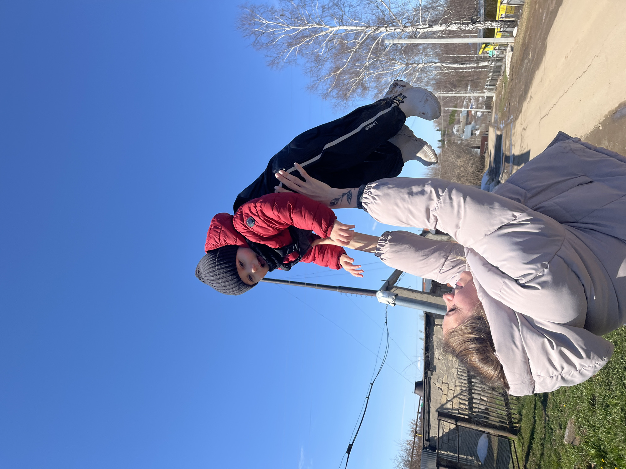 In Фоки, Russia, a person in a light grey coat lies on their back in a grassy area, holding a child upside down in the air. The child wears a red jacket, a black hat, and black pants with white stripes. The sky is clear and blue. In the background, there is a concrete path, a few trees, and some buildings.