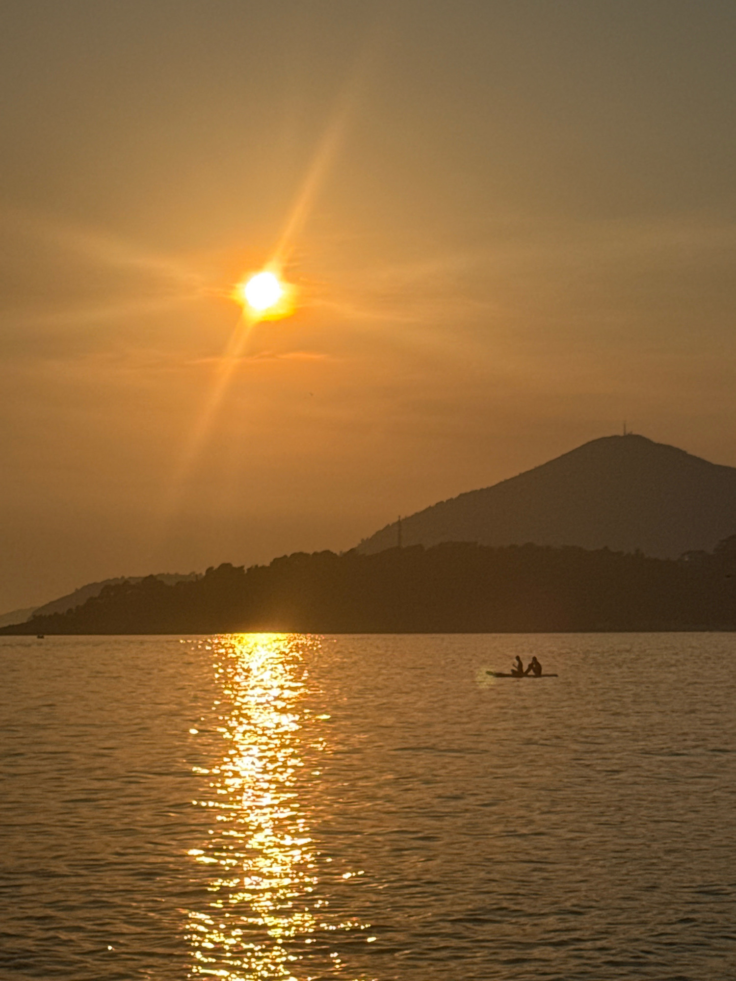 The image depicts a serene sunset over the Adriatic Sea in Rafailovići, Montenegro. The sun, a bright orb with distinct rays, is positioned in the upper left quadrant, casting a warm golden glow across the sky and water. A silhouetted mountain range forms the backdrop, with a prominent peak featuring a telecommunications tower. In the middle ground, a narrow strip of land, likely a coastline, is visible in silhouette. The water's surface is rippled, reflecting the sun's light in a shimmering path. A small raft with two silhouetted figures, appearing to be engaged in recreational activity, floats in the water, positioned to the right of the sun's reflection. The overall atmosphere is tranquil and picturesque.