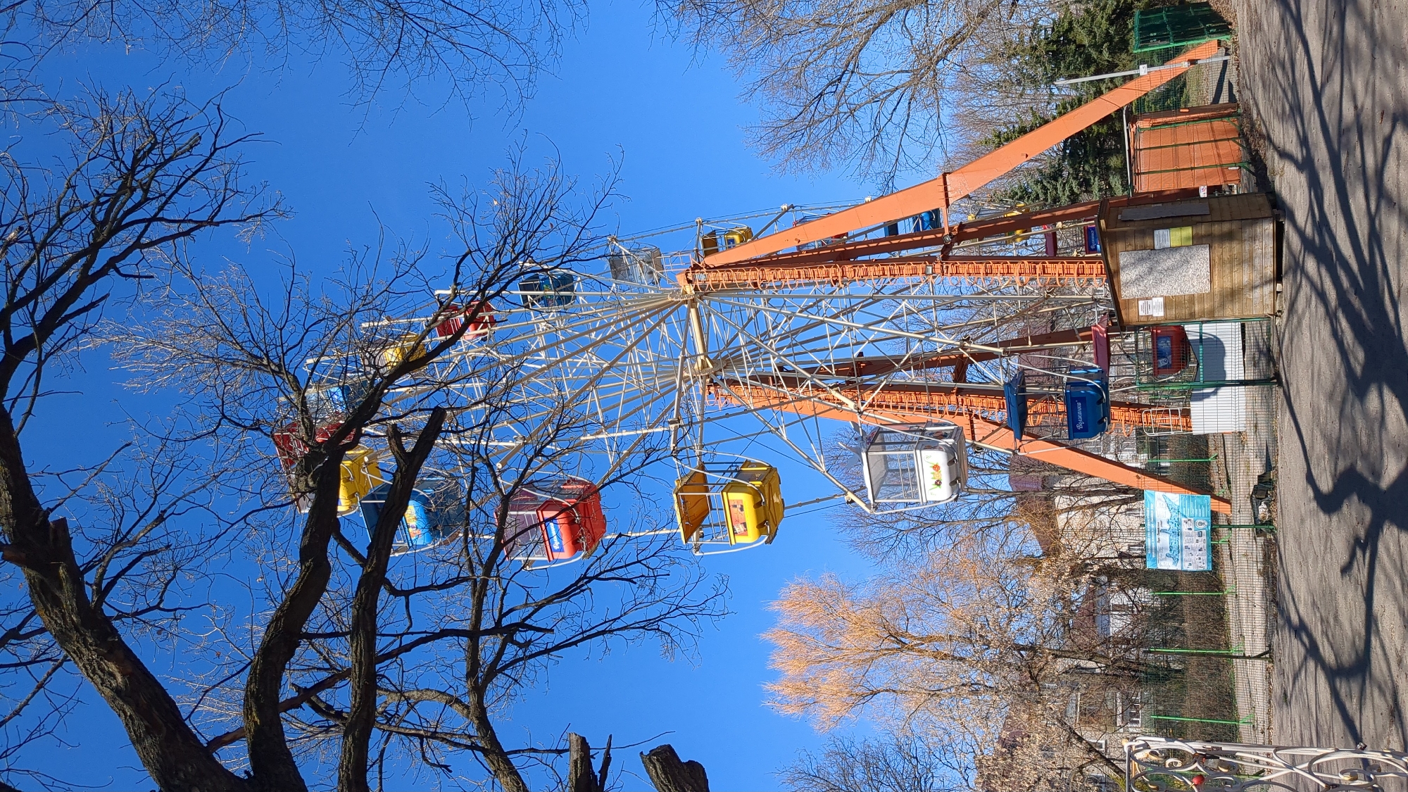 The image captures a Ferris wheel located in what appears to be a park in Ulyanovsk, Russia. The Ferris wheel has multiple colorful gondolas (red, yellow, blue, and white) and an orange-colored support structure. The image is taken from a low angle, looking up at the Ferris wheel against a clear, bright blue sky. Bare tree branches are visible in the foreground, partially obscuring the Ferris wheel. Adjacent to the base of the Ferris wheel is a small building and a sign with information displayed on it. A paved area runs alongside the Ferris wheel, indicating a pathway within the park. The overall environment suggests an urban recreational space on a sunny day.