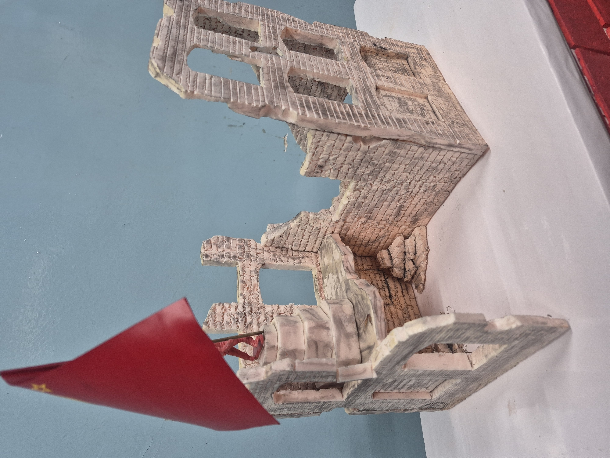 A model of a partially destroyed building with exposed brick-like material is affixed to a white wall, with a light blue wall in the background. A red flag is positioned to the lower left of the model, appearing to be planted within or near it. The location is Chaikovsky, Russia.