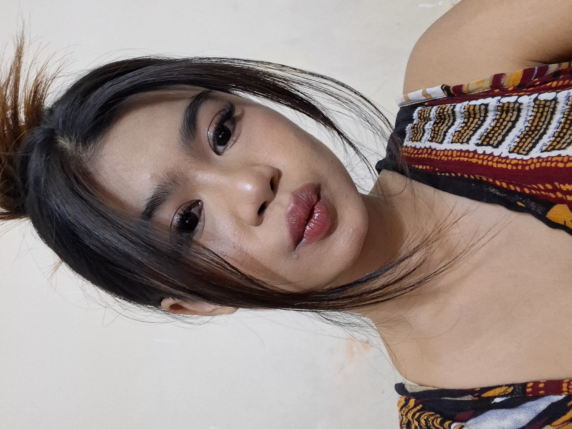 This is a close-up, waist-up, portrait-style photograph of a young woman with dark hair pulled up loosely, with some strands falling around her face. She is wearing an off-the-shoulder top with a vibrant, patterned fabric featuring geometric designs in red, yellow, white, and black. Her makeup is noticeable, with well-defined eyebrows, prominent eyelashes, and glossy, reddish-pink lipstick. The lighting suggests an indoor setting, possibly with artificial light. The background is a plain, light-colored wall. The woman's expression is a direct gaze into the camera, with a slightly parted mouth, conveying a sense of self-awareness or posing. The overall tone of the image is casual and personal, likely a selfie. Given the location context of Kuta, Indonesia, the image might be from a tropical or warm climate setting. There is no visible text in the image.